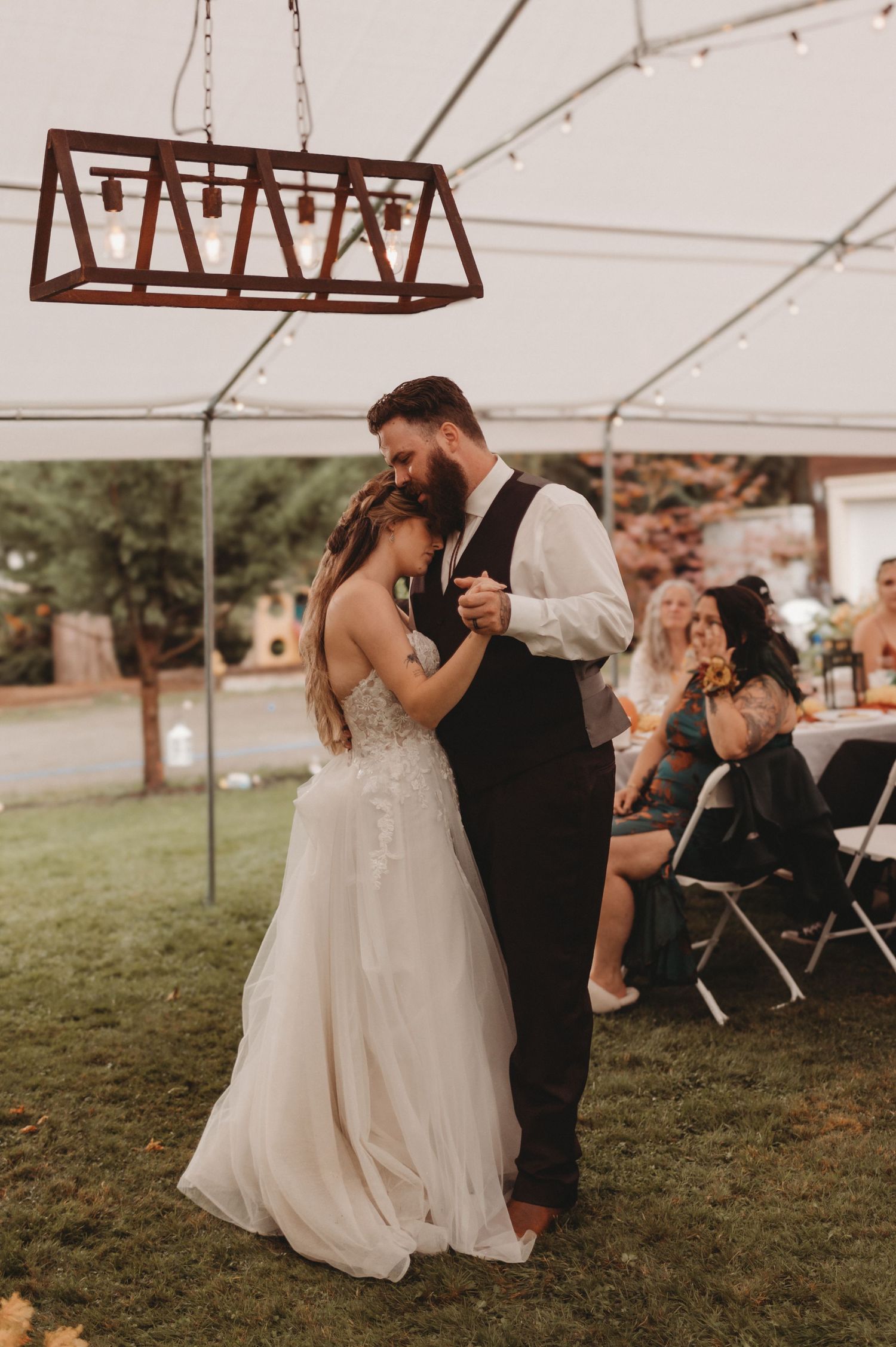 A bride and bearded groom share their first dance under a tent with string lights at their reception.