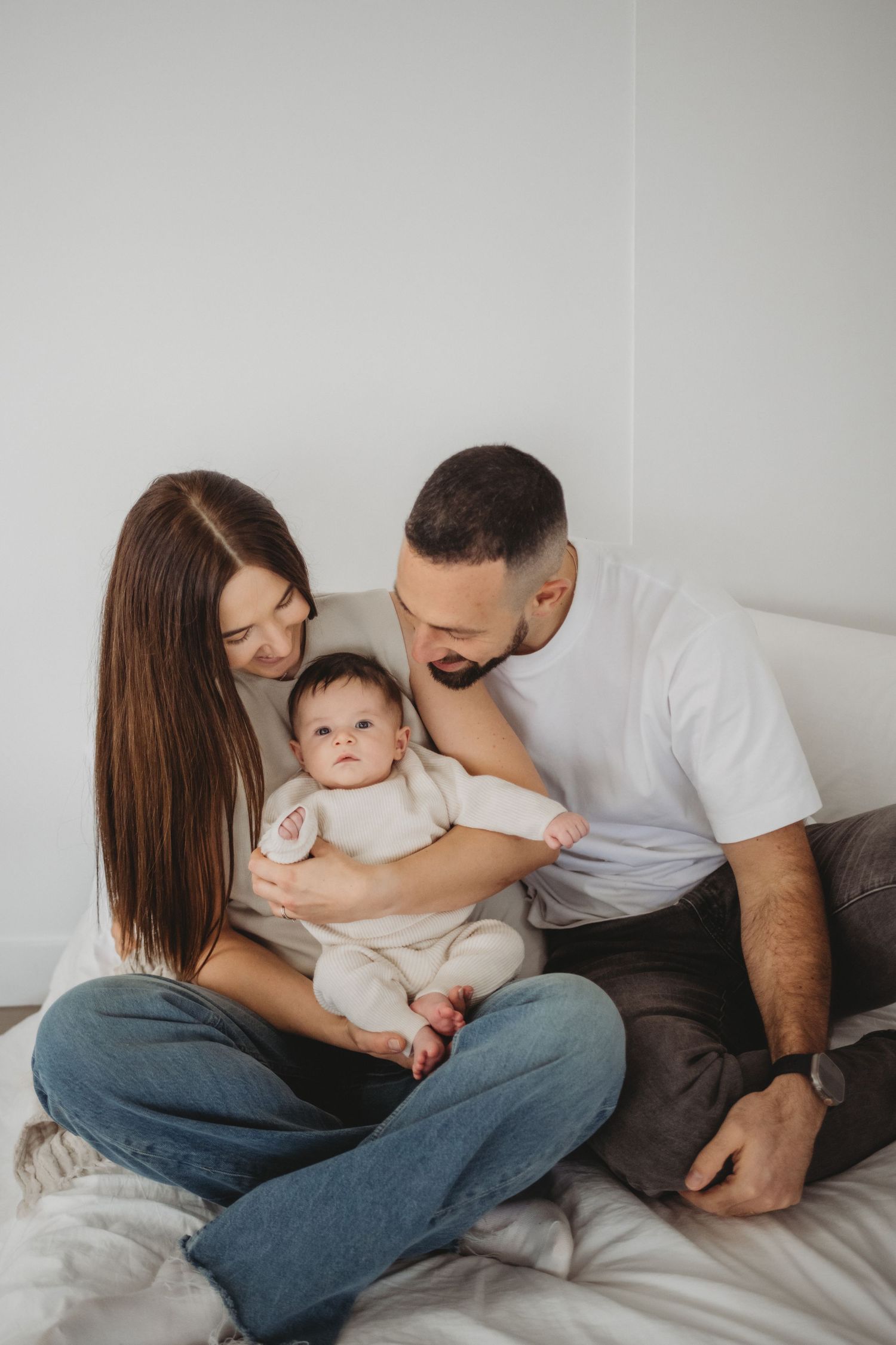 Loving parents on a bed leaning in close to their alert newborn baby dressed in white.