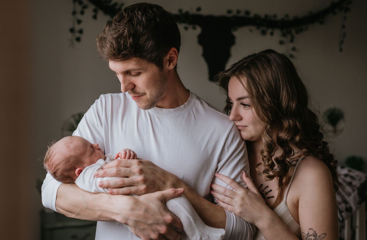 New father in white shirt holding newborn baby with mother beside him in cozy nursery.