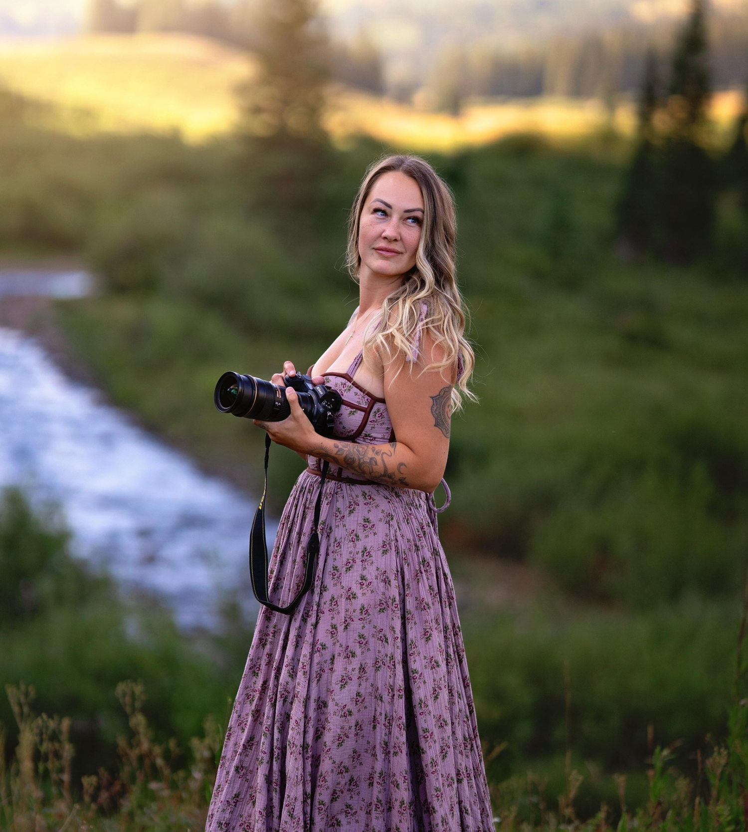 Blonde female photographer in floral dress holding camera near mountain stream at golden hour.