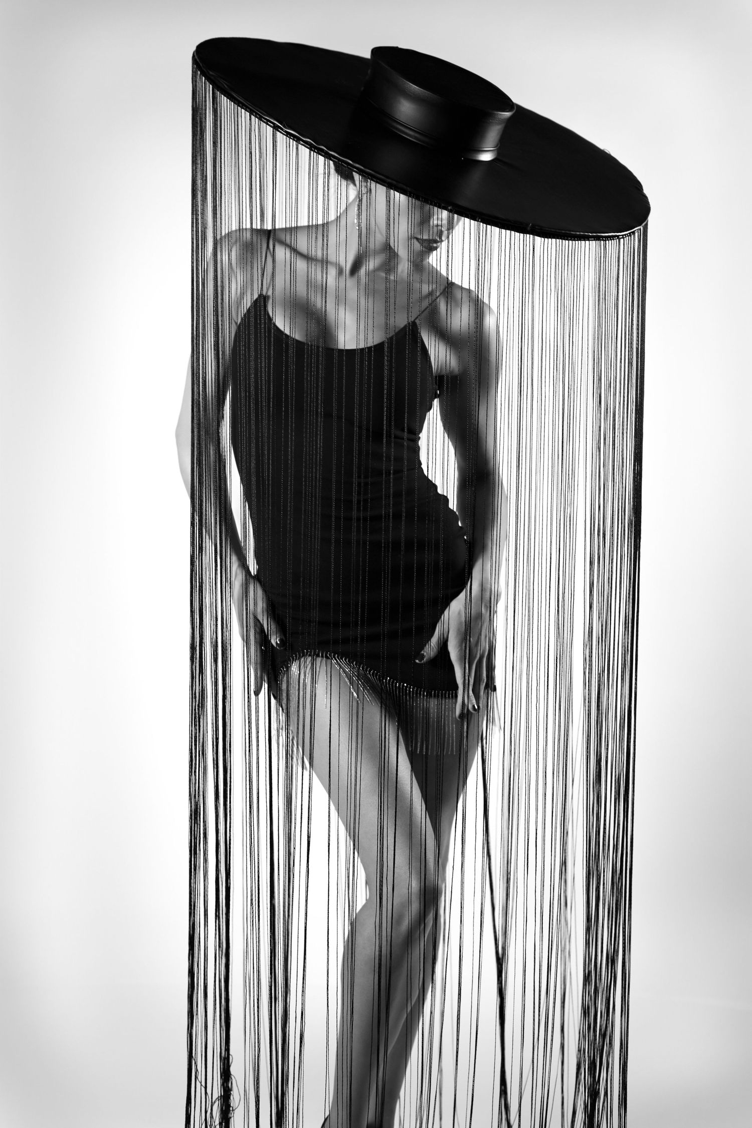 Black and white fine art studio portrait of the model, posing elegantly, looking down, wearing a black fringe hat.