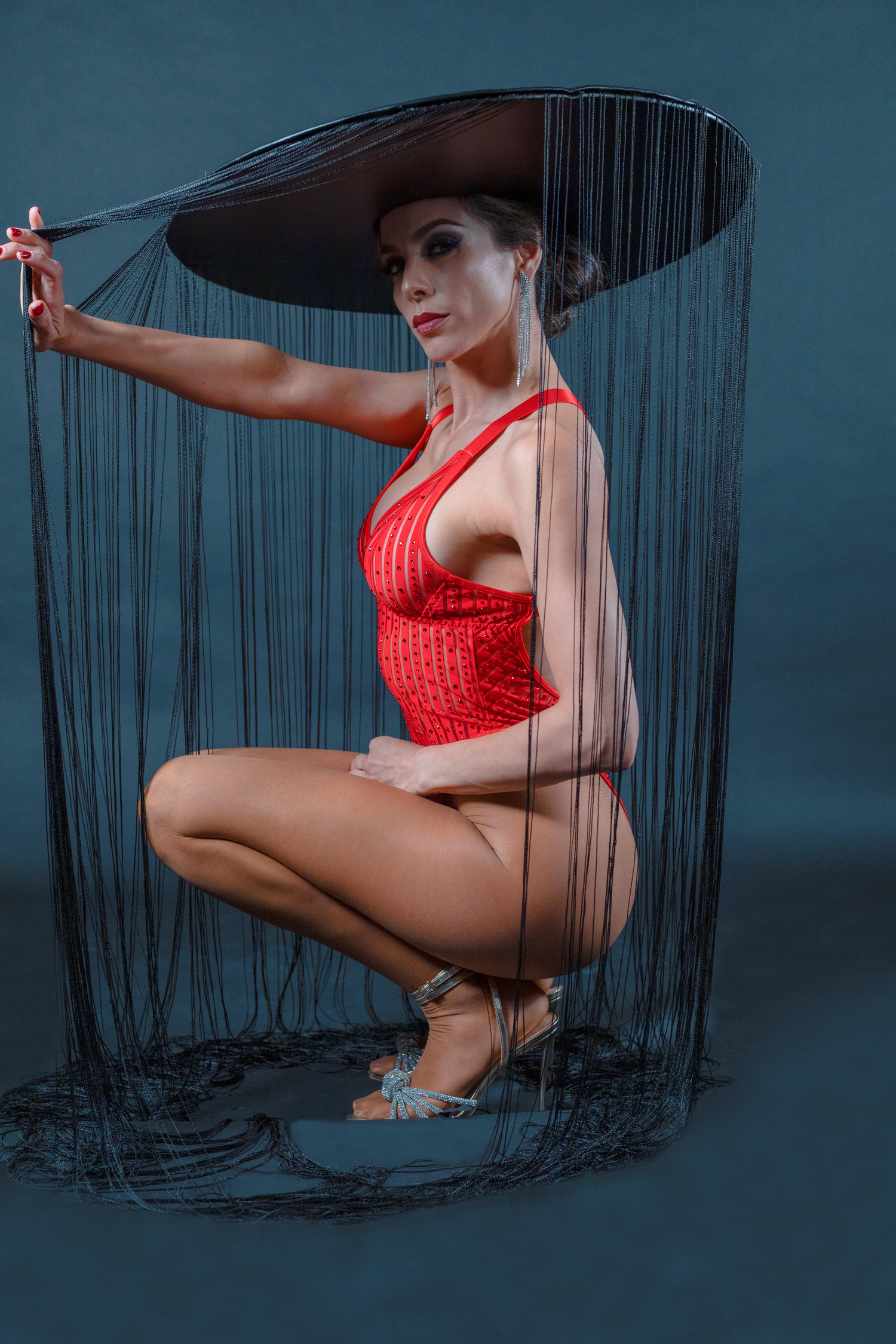 Studio portrait of the model crouching down with a red lace dress, and a black fringe hat, looking at the camera.