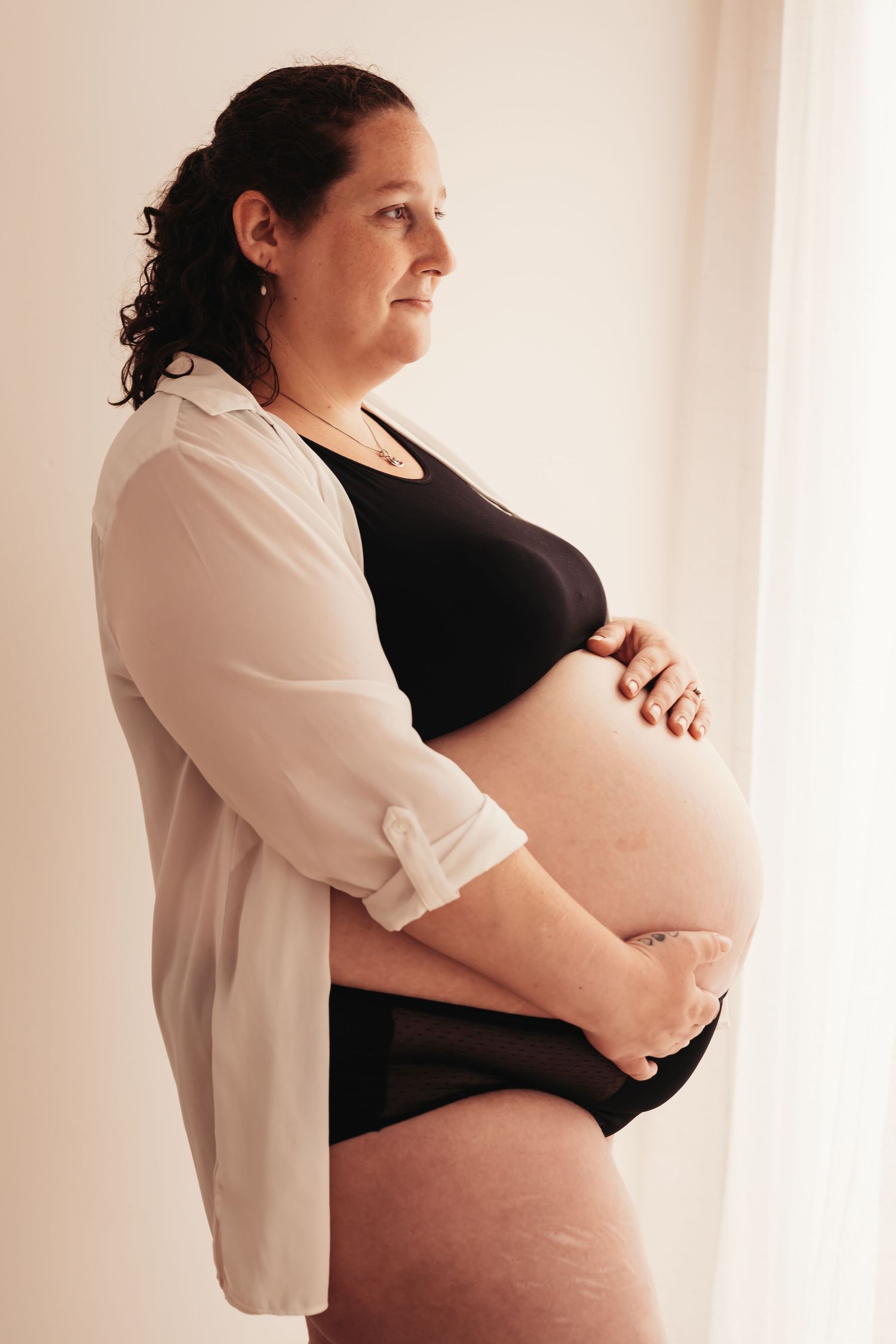 Maternity photo showing expectant mother in white cardigan against bright window.