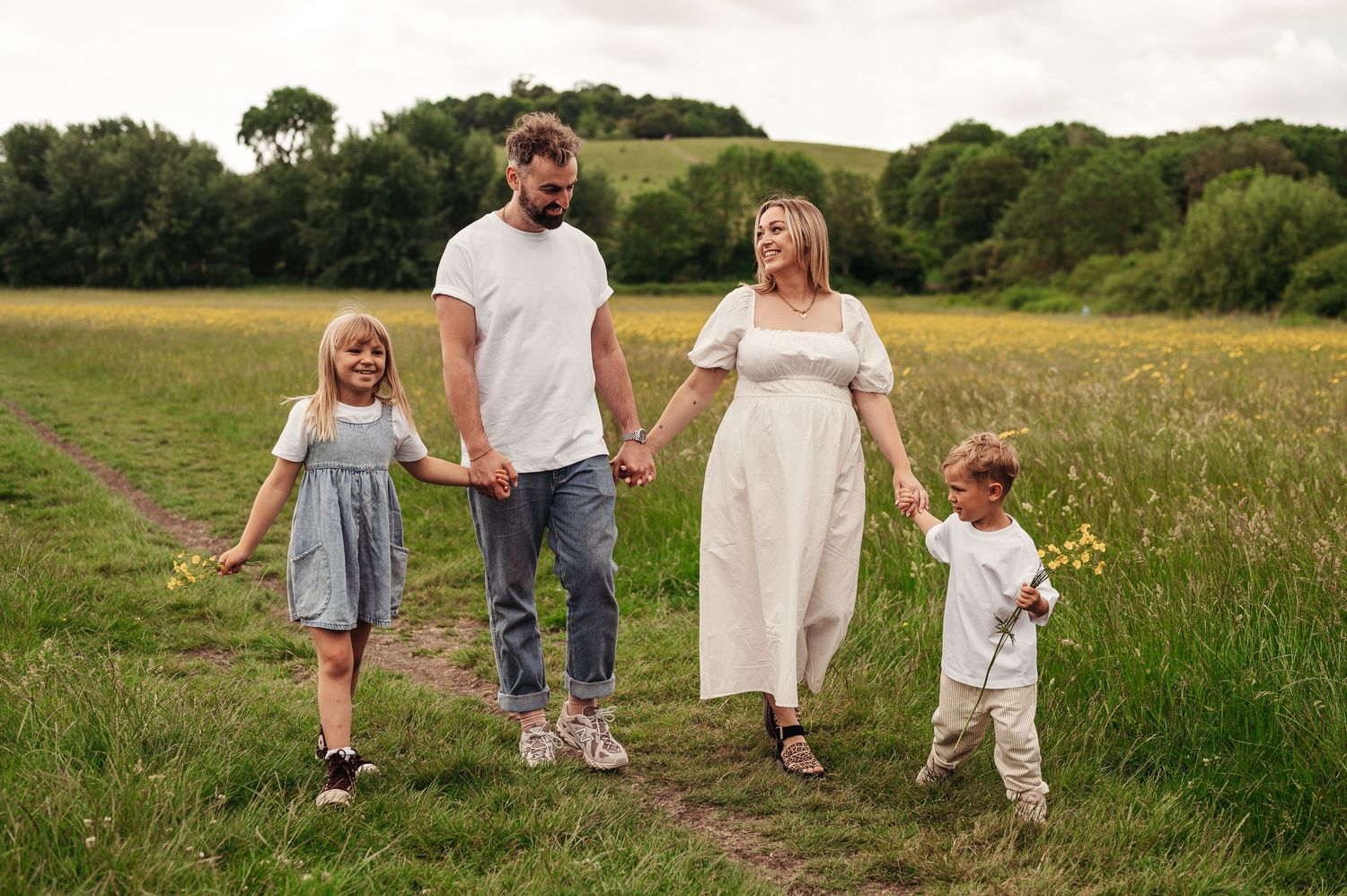 A family walks together holding hands on a grassy path through a summer meadow during a sunset photoshoot.