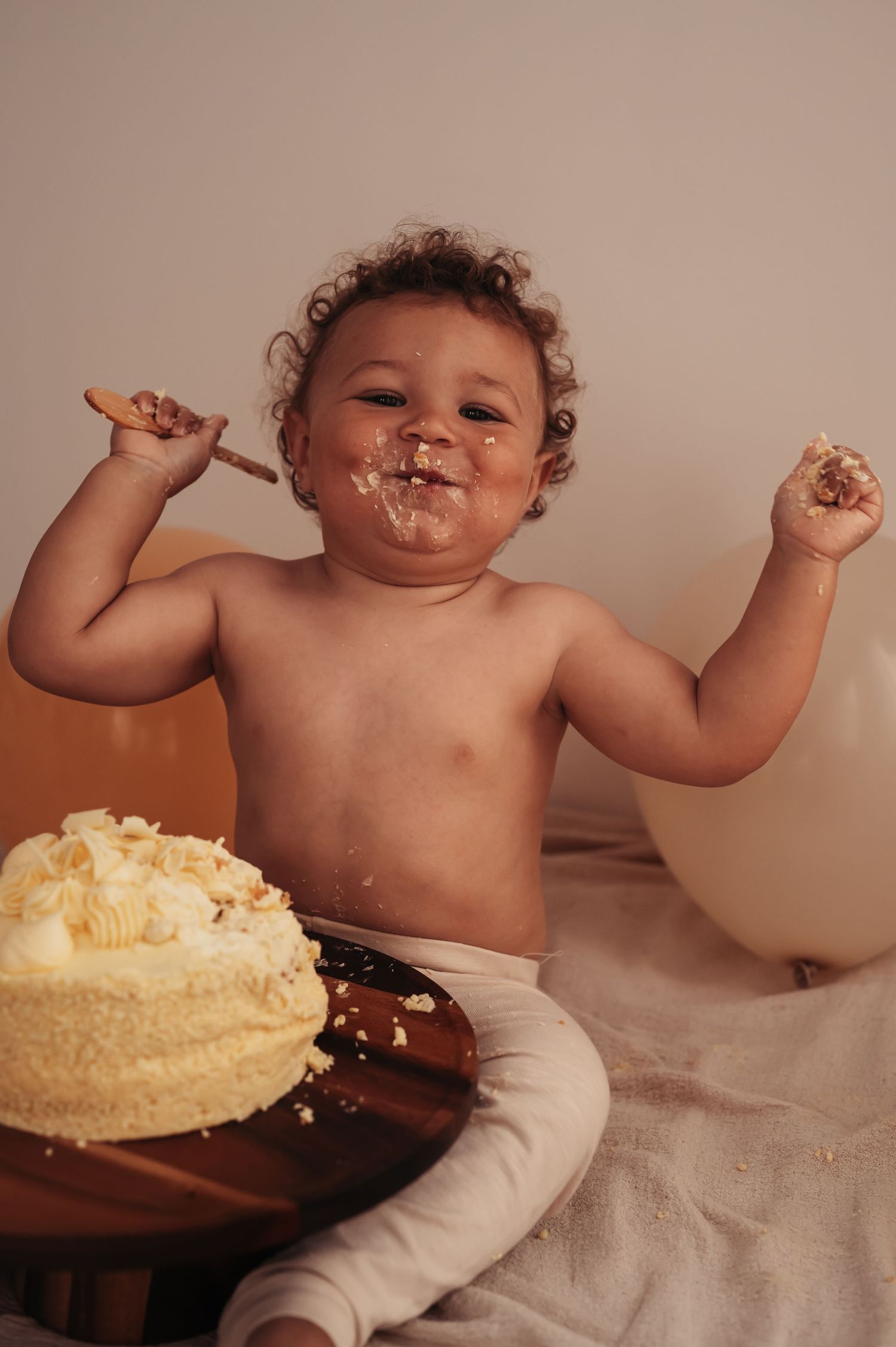 Joyful toddler covered in cake frosting raising arms in celebration during cake smash session.