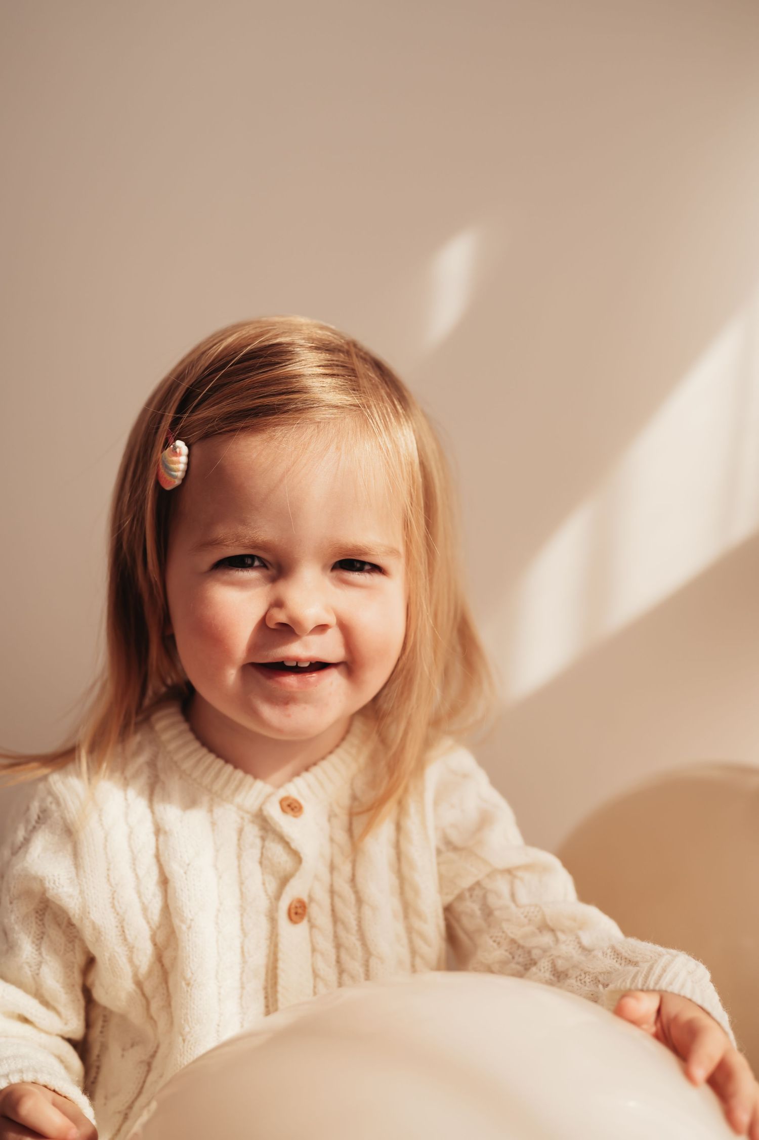 Smiling toddler in cream cable knit sweater poses against neutral wall with soft lighting.