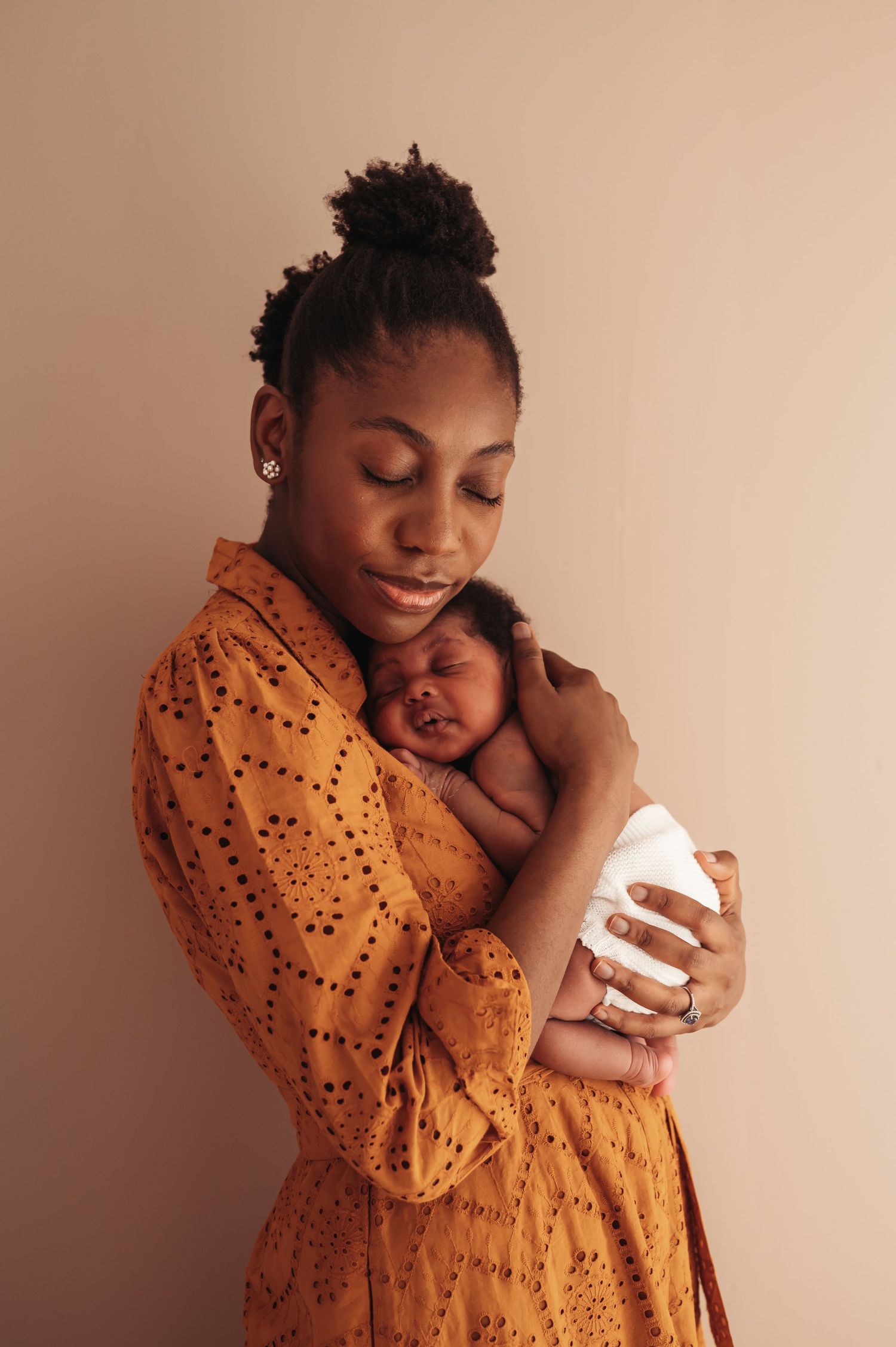 A young Black mother in an orange dress tenderly holds her swaddled newborn baby close to her chest.