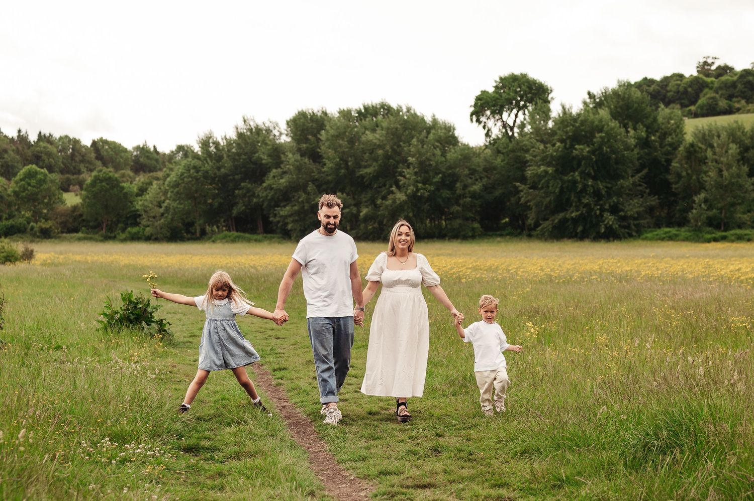 A family walks together along a dirt path through a sunny meadow surrounded by green trees in summer.