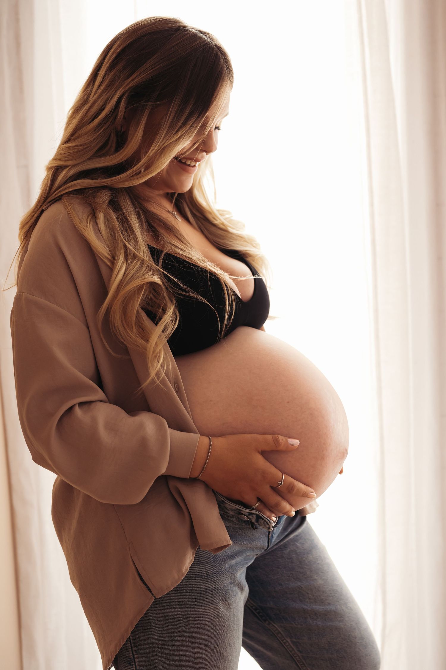 Pregnant woman in black bralette, beige blazer, and jeans cradling her bare baby bump while smiling downward near white curtains.