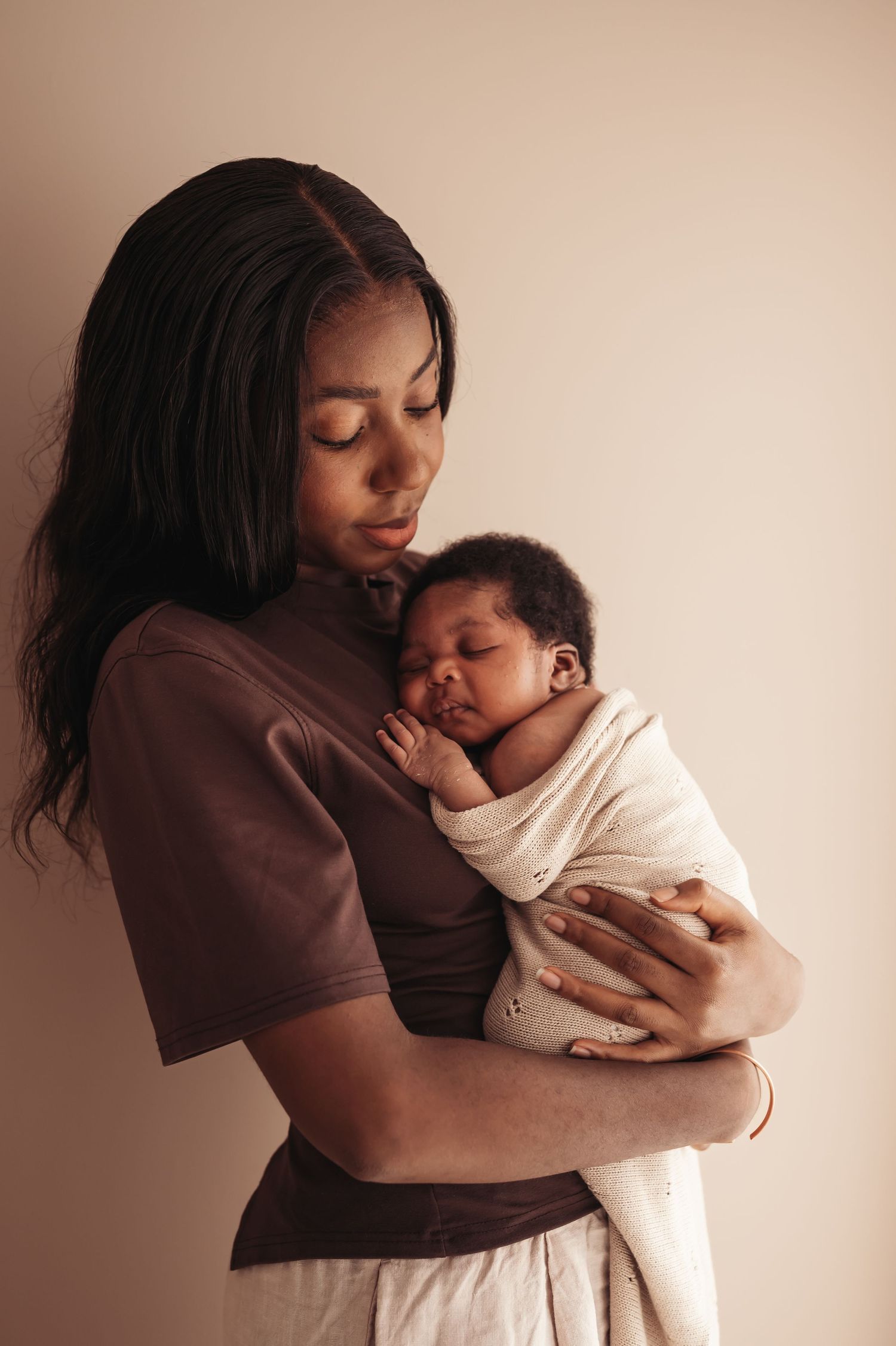A young Black mother tenderly holds her sleeping newborn baby wrapped in white cloth against her chest.