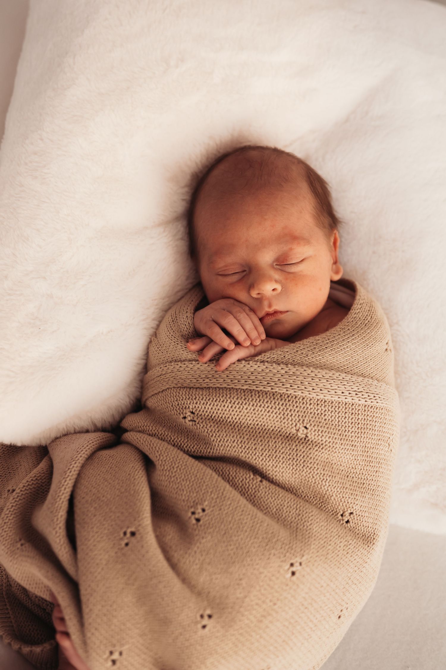 Sleeping newborn swaddled in textured beige blanket on white surface.