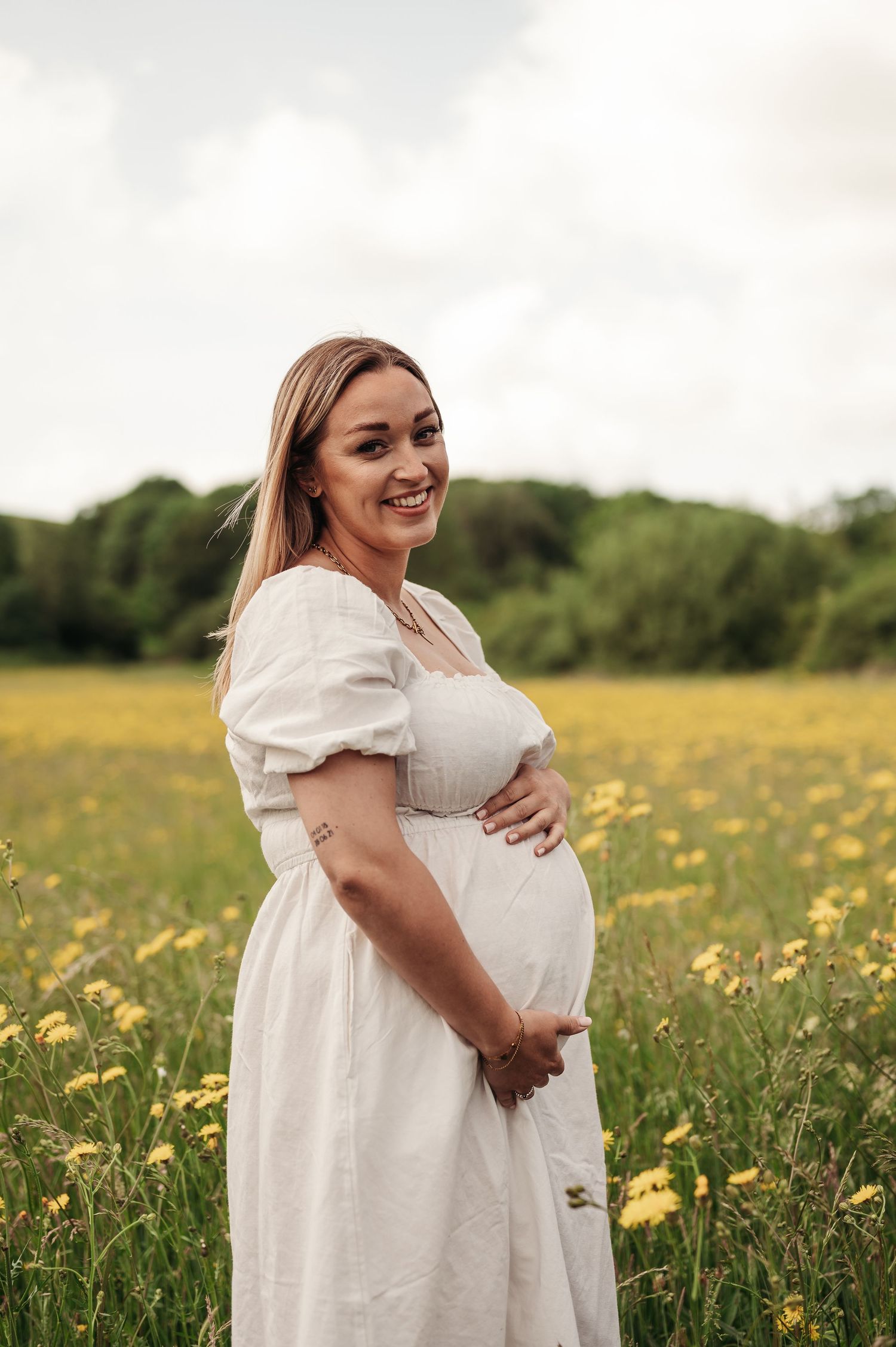 Expectant mother in white dress stands joyfully in wildflower meadow on bright summer day.