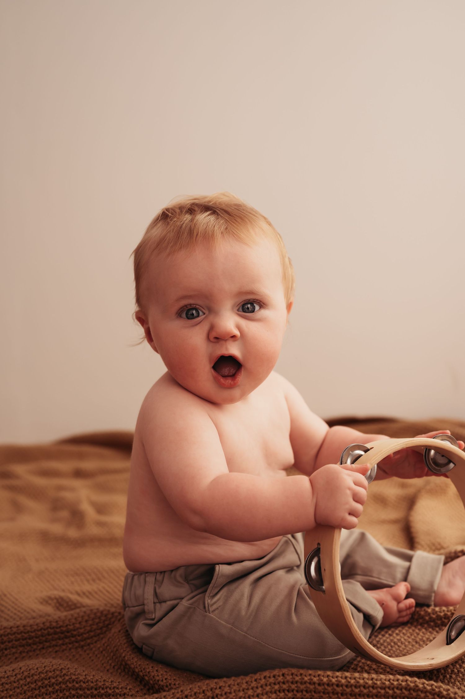 Happy baby in tan pants plays with toy while sitting on brown blanket.