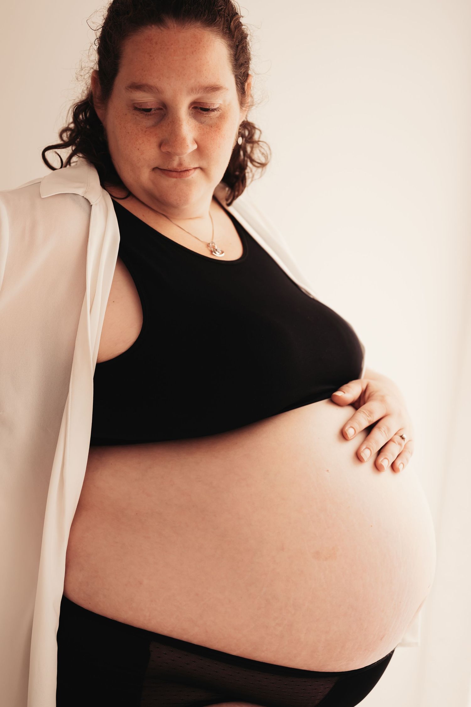Maternity photo showing a pregnant figure in black top and white cardigan against neutral wall.