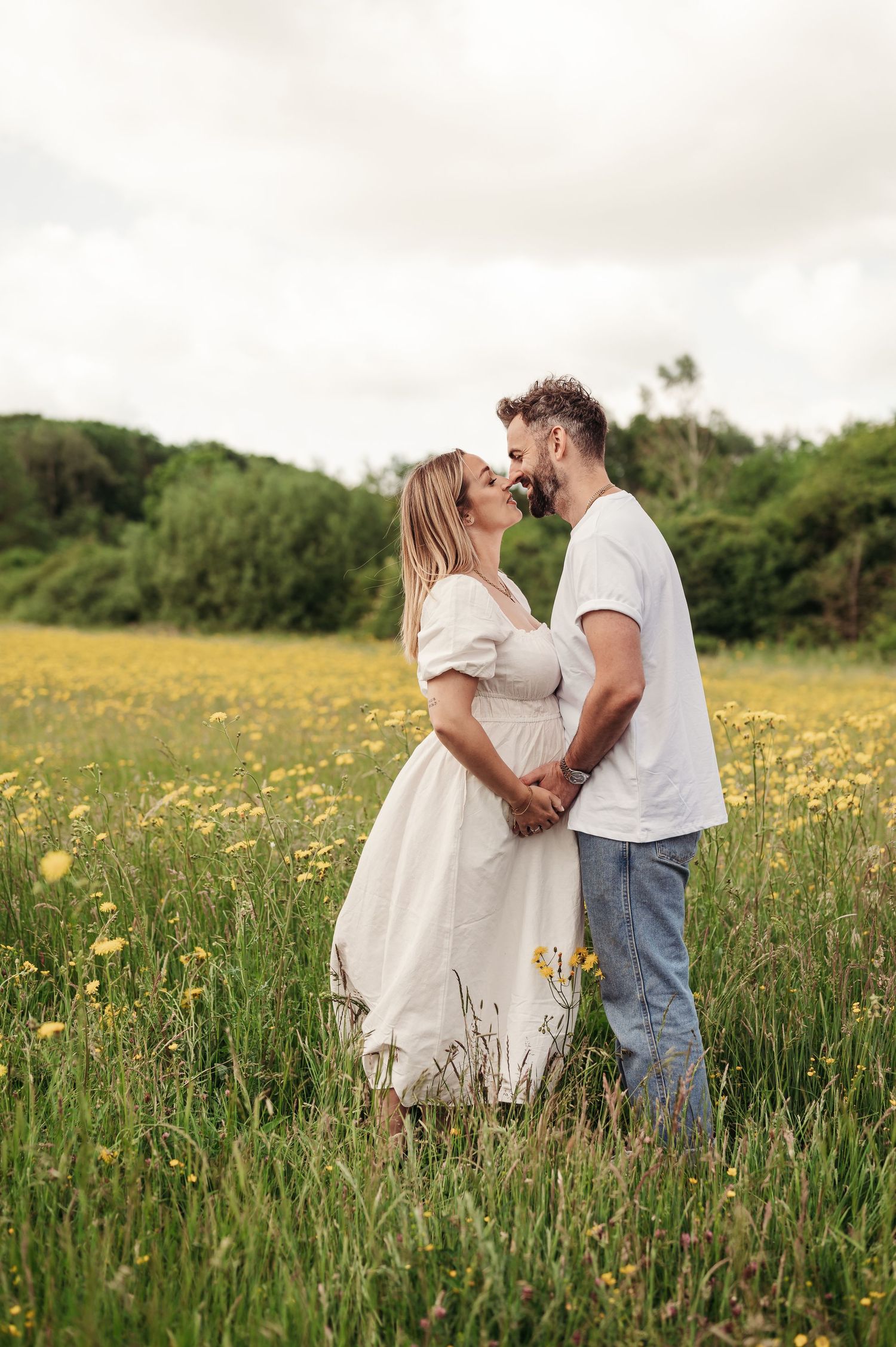 A couple embraces lovingly in a sunny meadow filled with yellow wildflowers and green grass on a cloudy day.