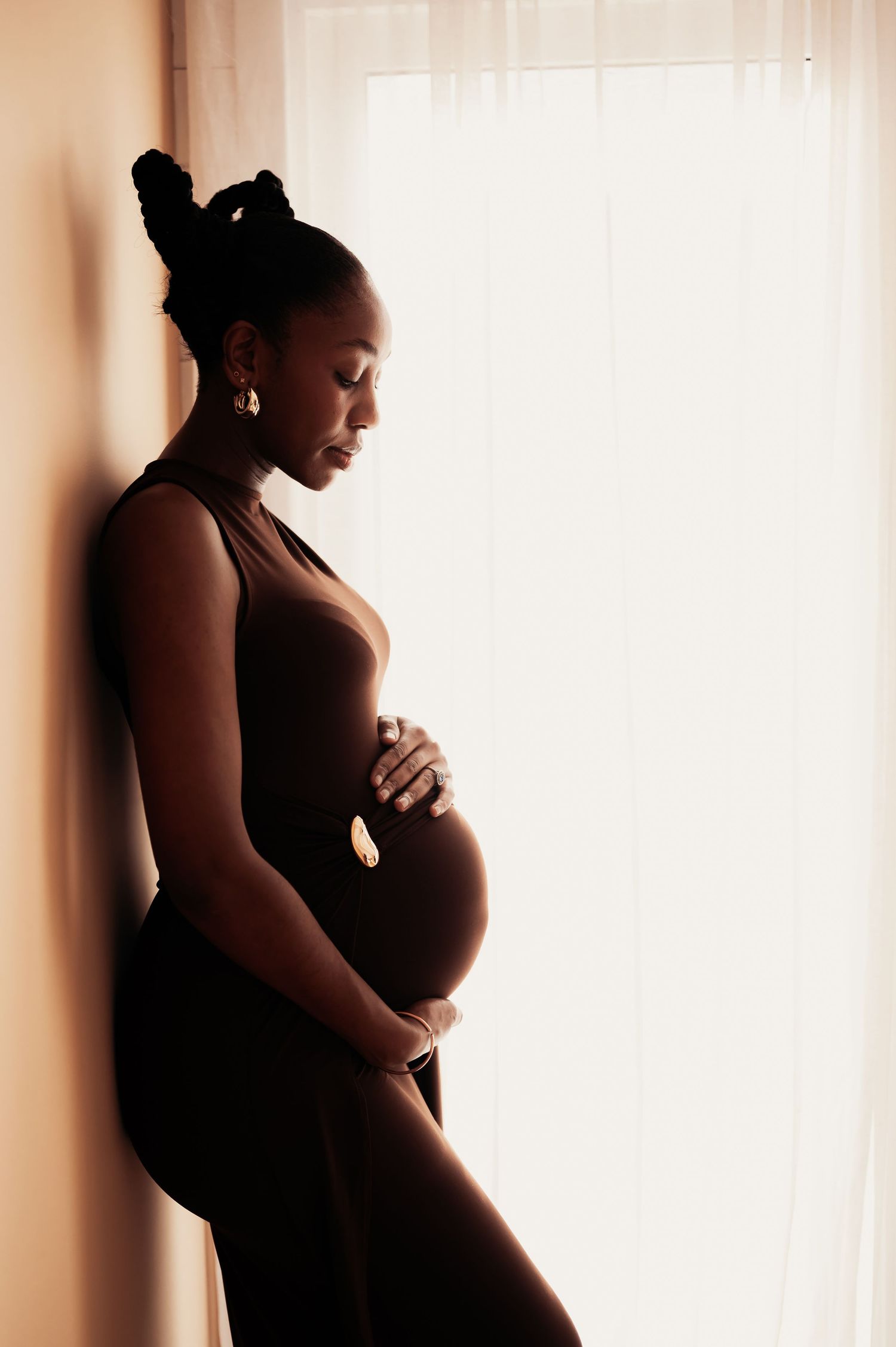 Pregnant Black woman in dark bodysuit cradling baby bump, standing by window in soft warm light, maternity photography.