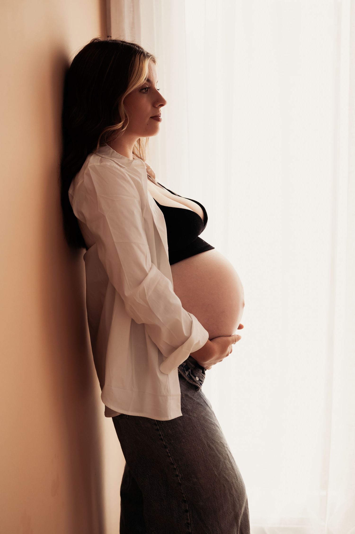 Pregnant woman in white shirt cradling baby bump, leaning against wall by window in soft natural light.