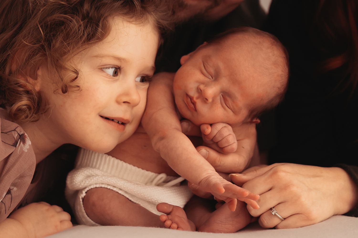 A curly-haired child smiles while looking at a sleeping newborn baby wrapped in beige.