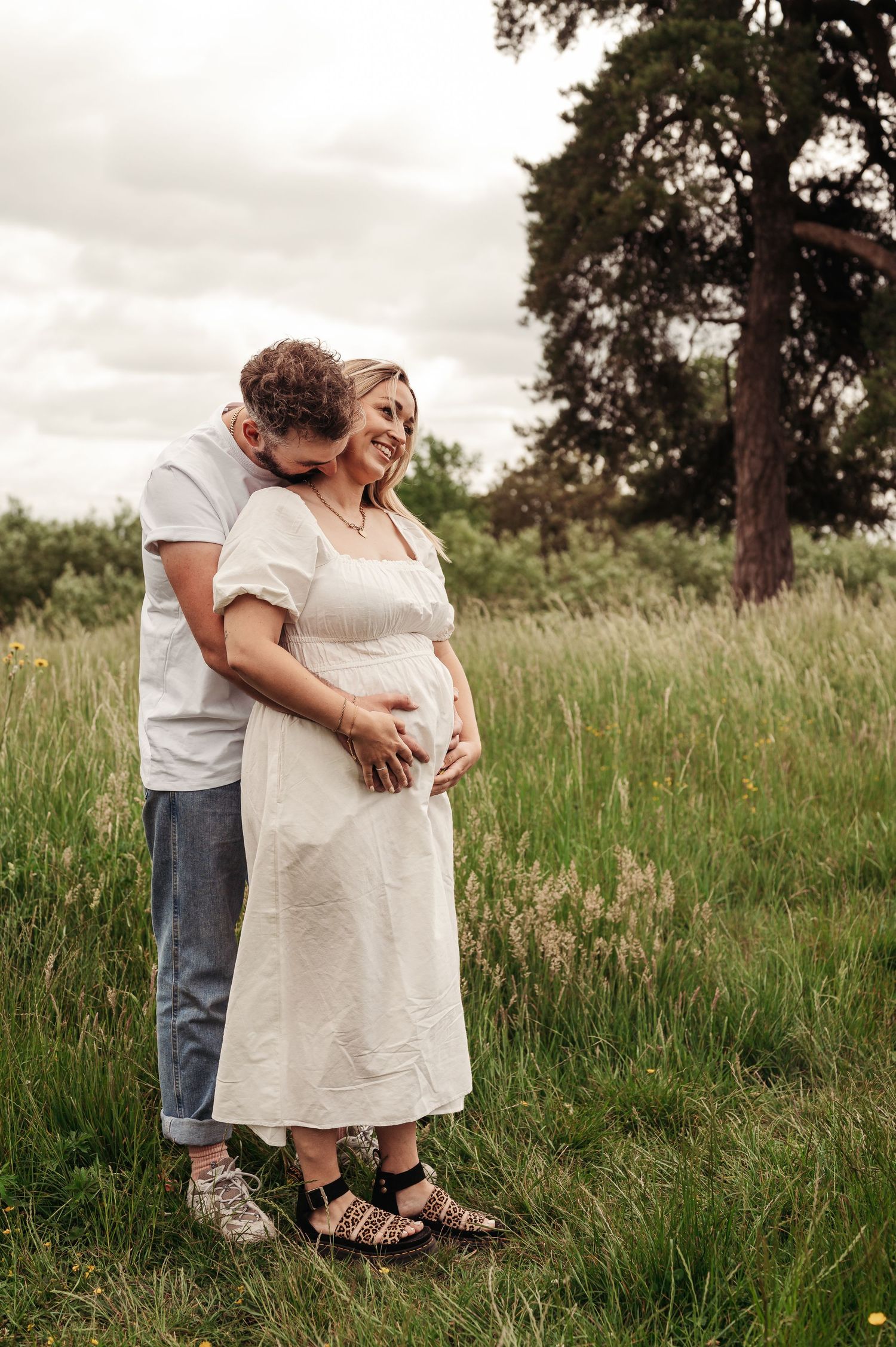 A couple embraces in tall grass with pine trees in the background on an overcast day.
