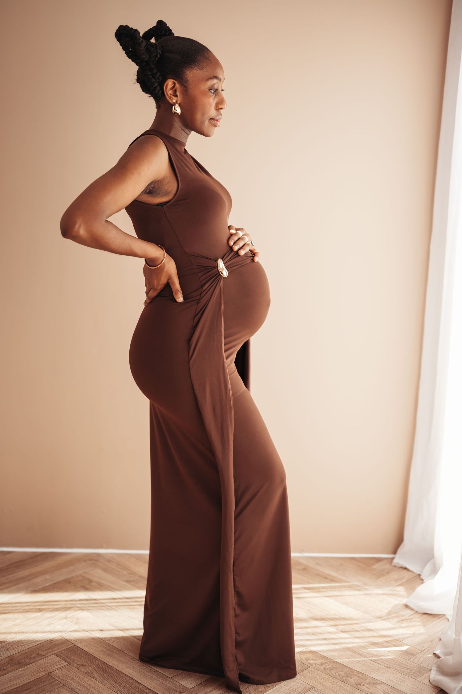 Pregnant woman in elegant brown maxi dress posing sideways, showcasing baby bump in a minimalist studio setting.
