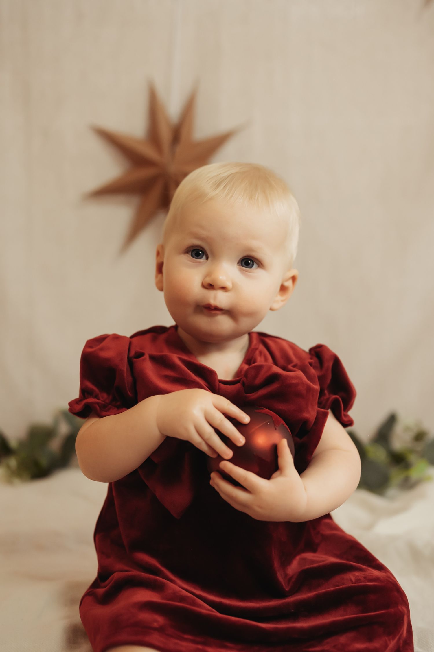 Baby in red velvet dress holds Christmas ornament with star decoration backdrop.
