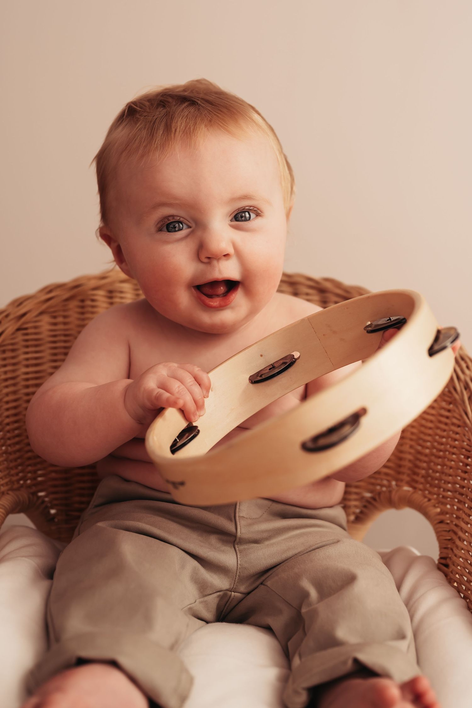 Happy baby holding wooden tambourine while sitting in wicker chair.
