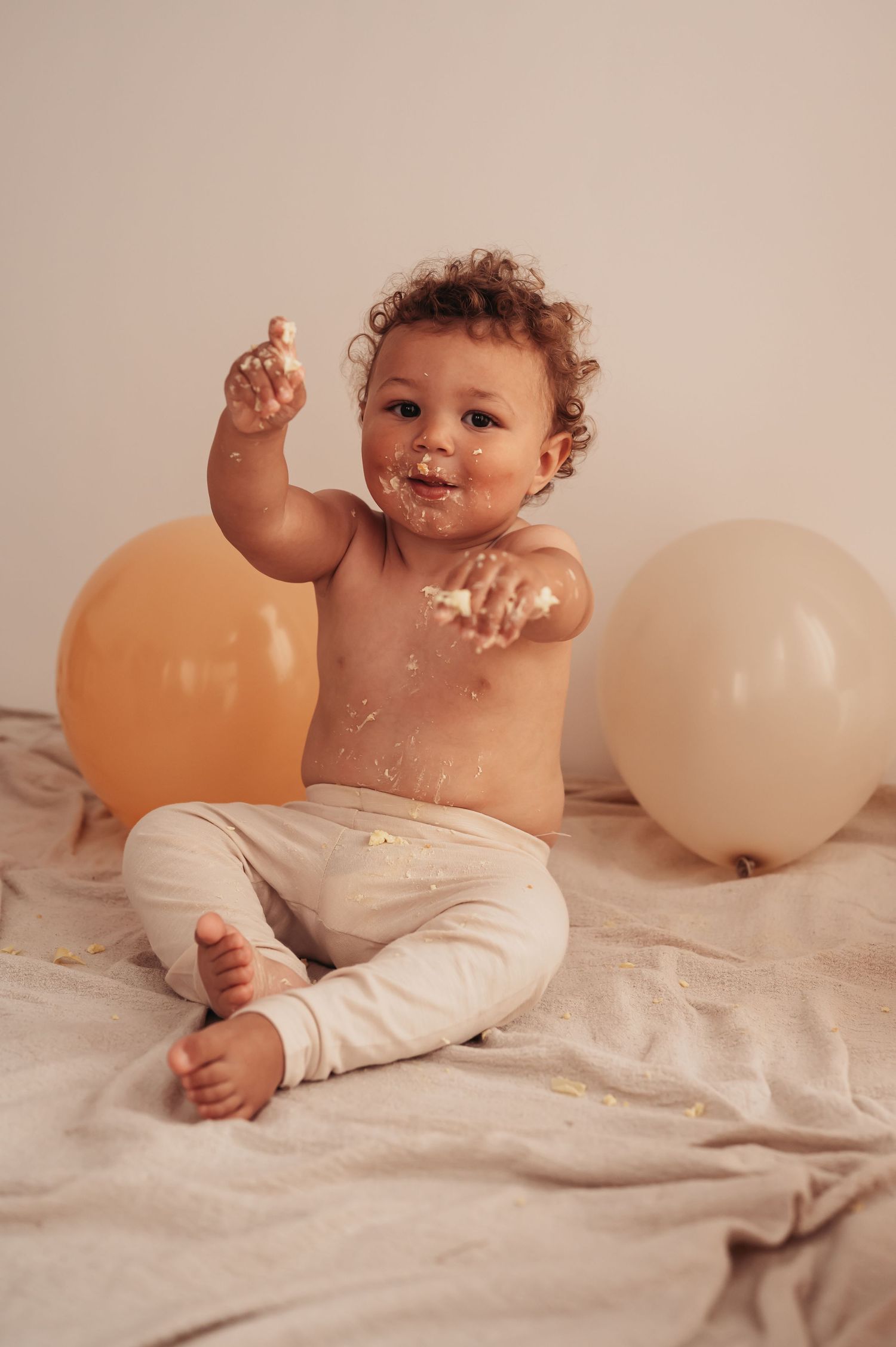 Happy baby raising frosting-covered hand during cake smash celebration with balloons.