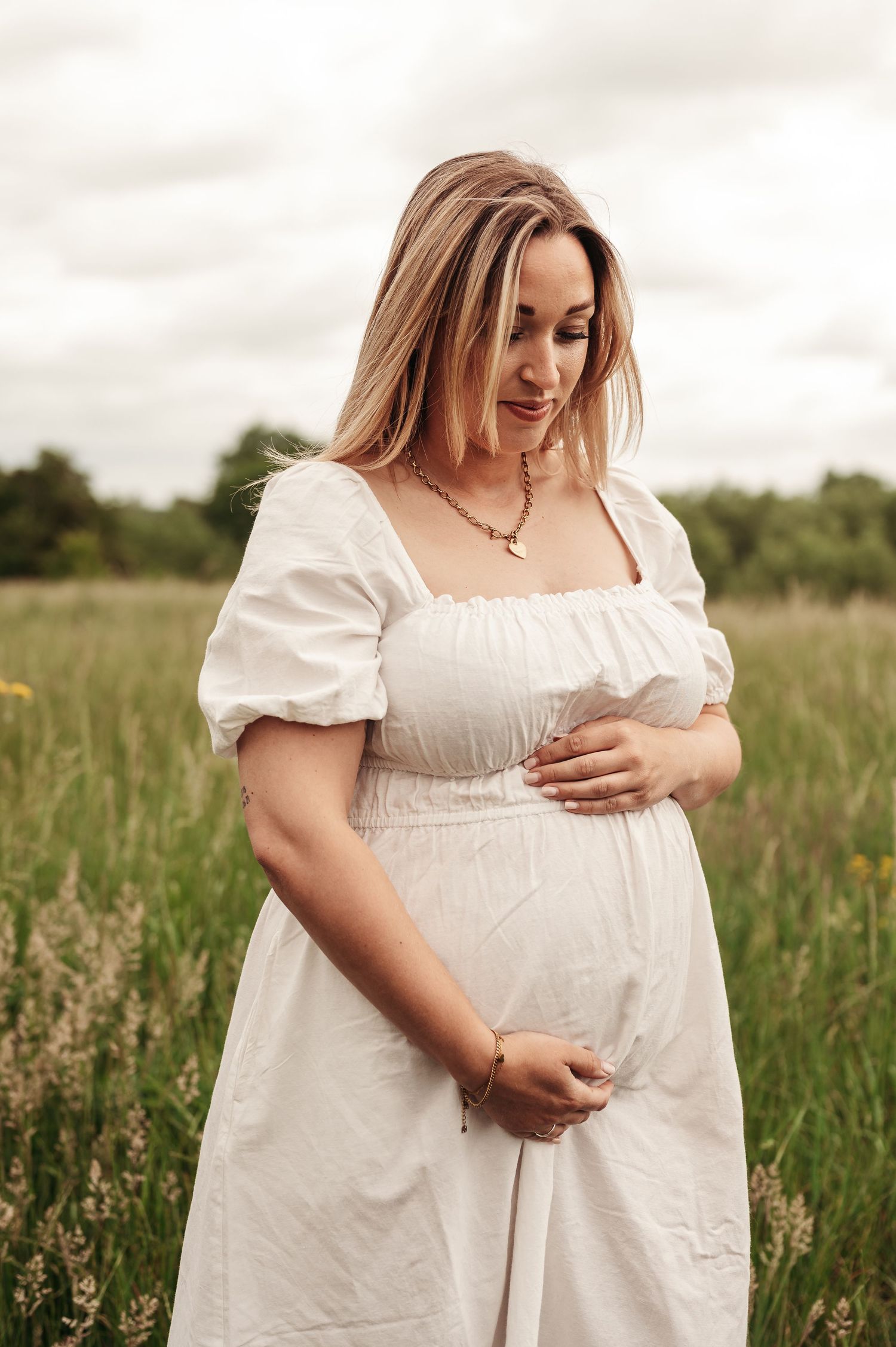 A woman in a white prairie dress stands in a sunny meadow with wildflowers, gently cradling her pregnant belly.