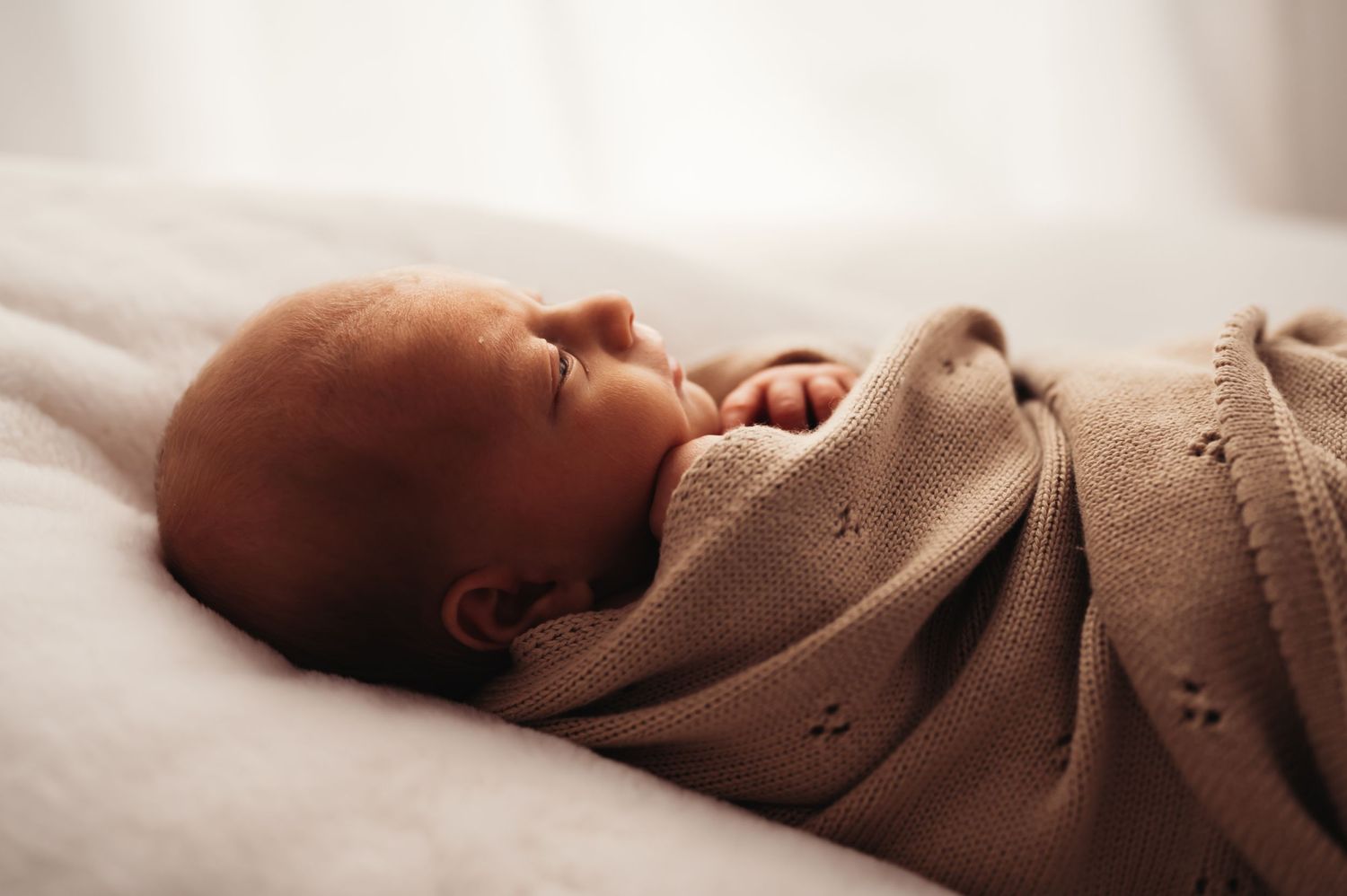 Side view of sleeping newborn wrapped in tan knit blanket on white bedding.