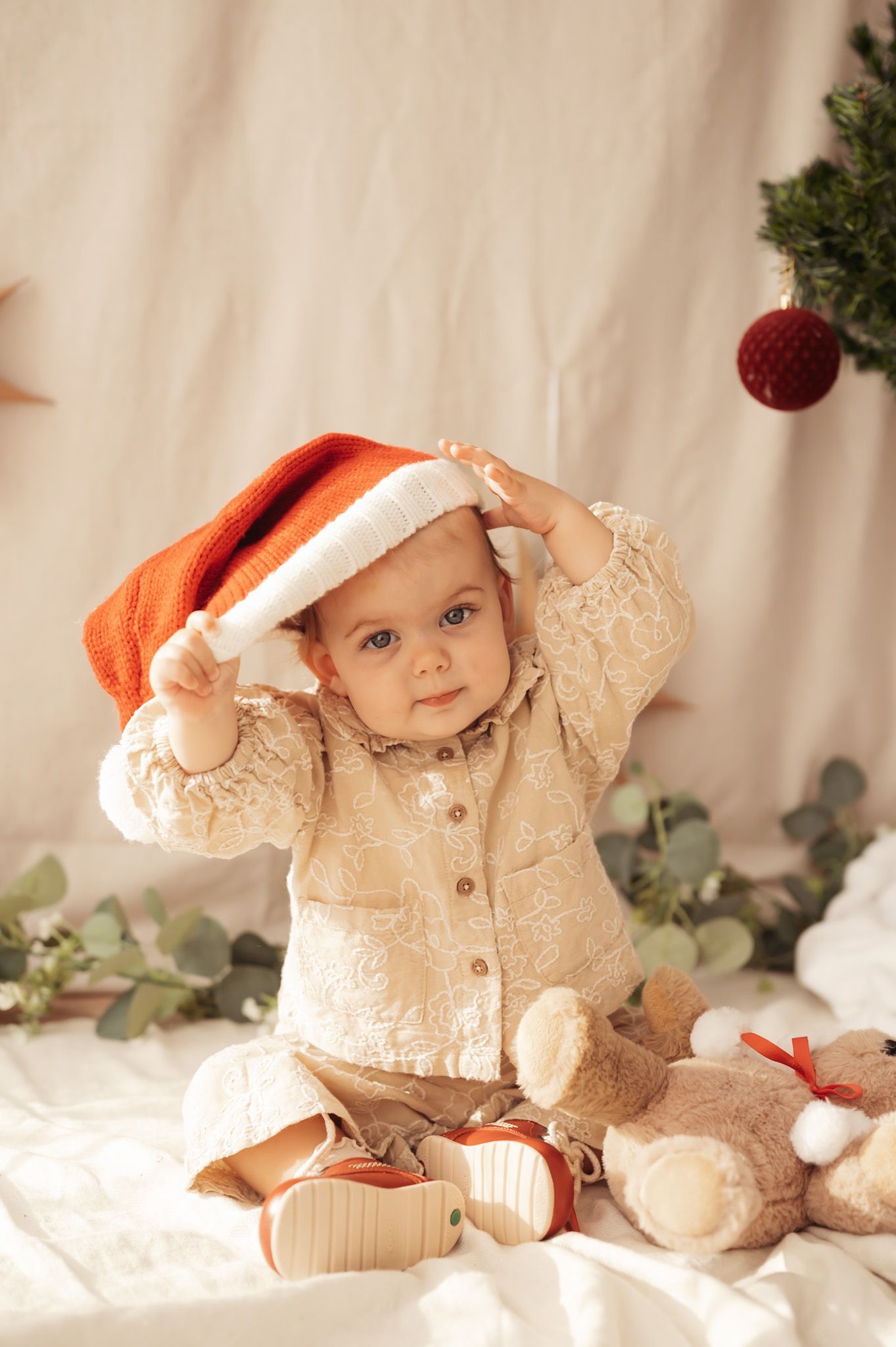 Adorable baby in Santa hat and cream outfit sits with teddy bear near Christmas tree decorations.