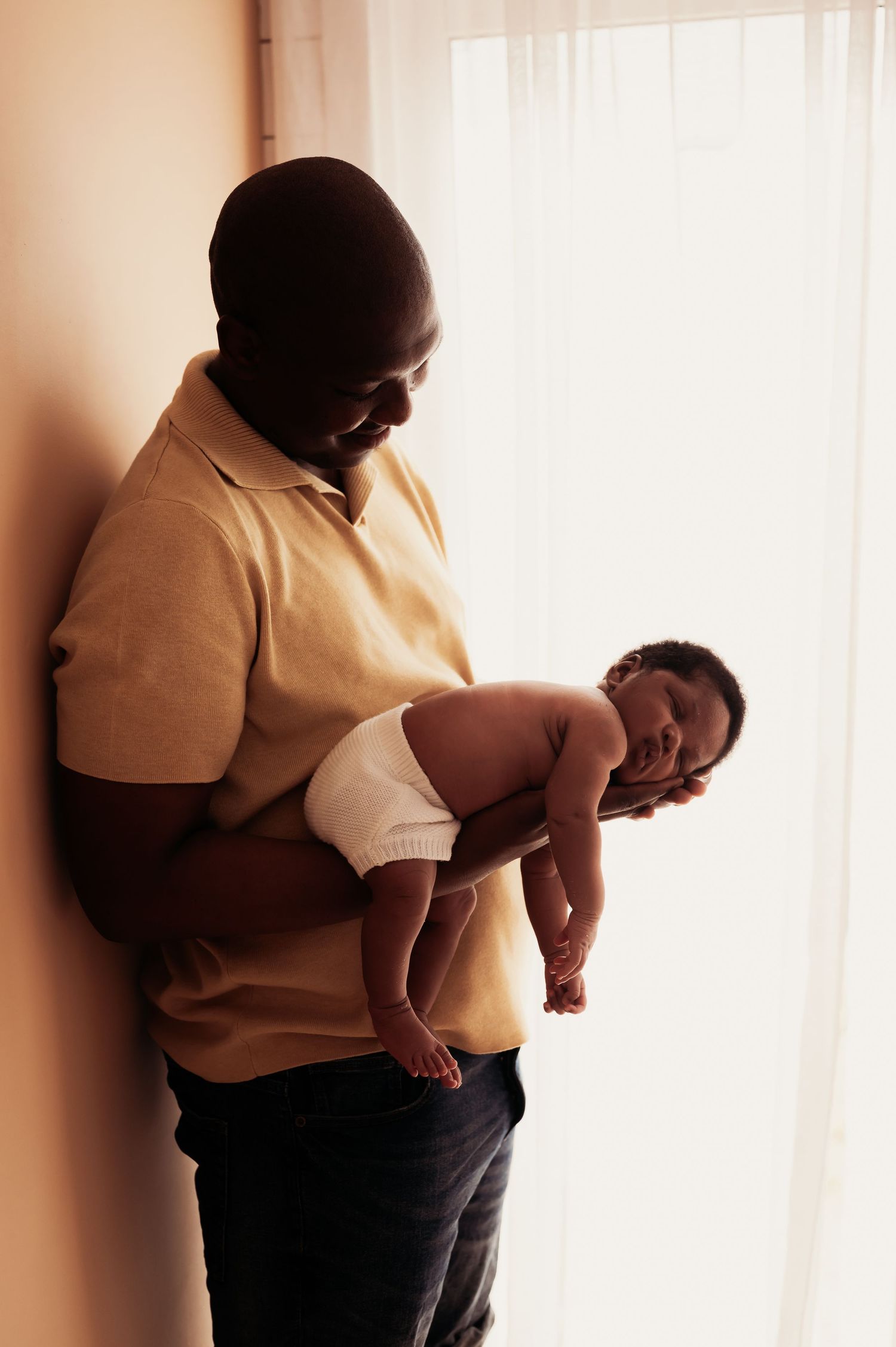 A father in a yellow polo shirt tenderly holds his sleeping newborn baby near a bright window.