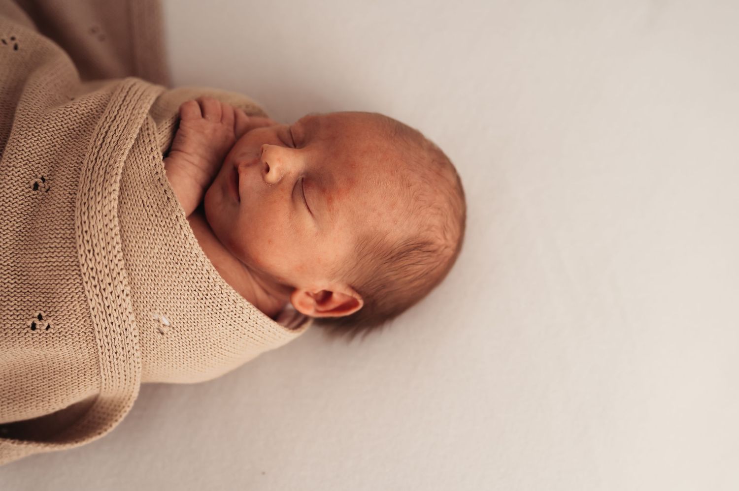 Peaceful newborn baby swaddled in a soft beige knit blanket, sleeping soundly on a white surface.