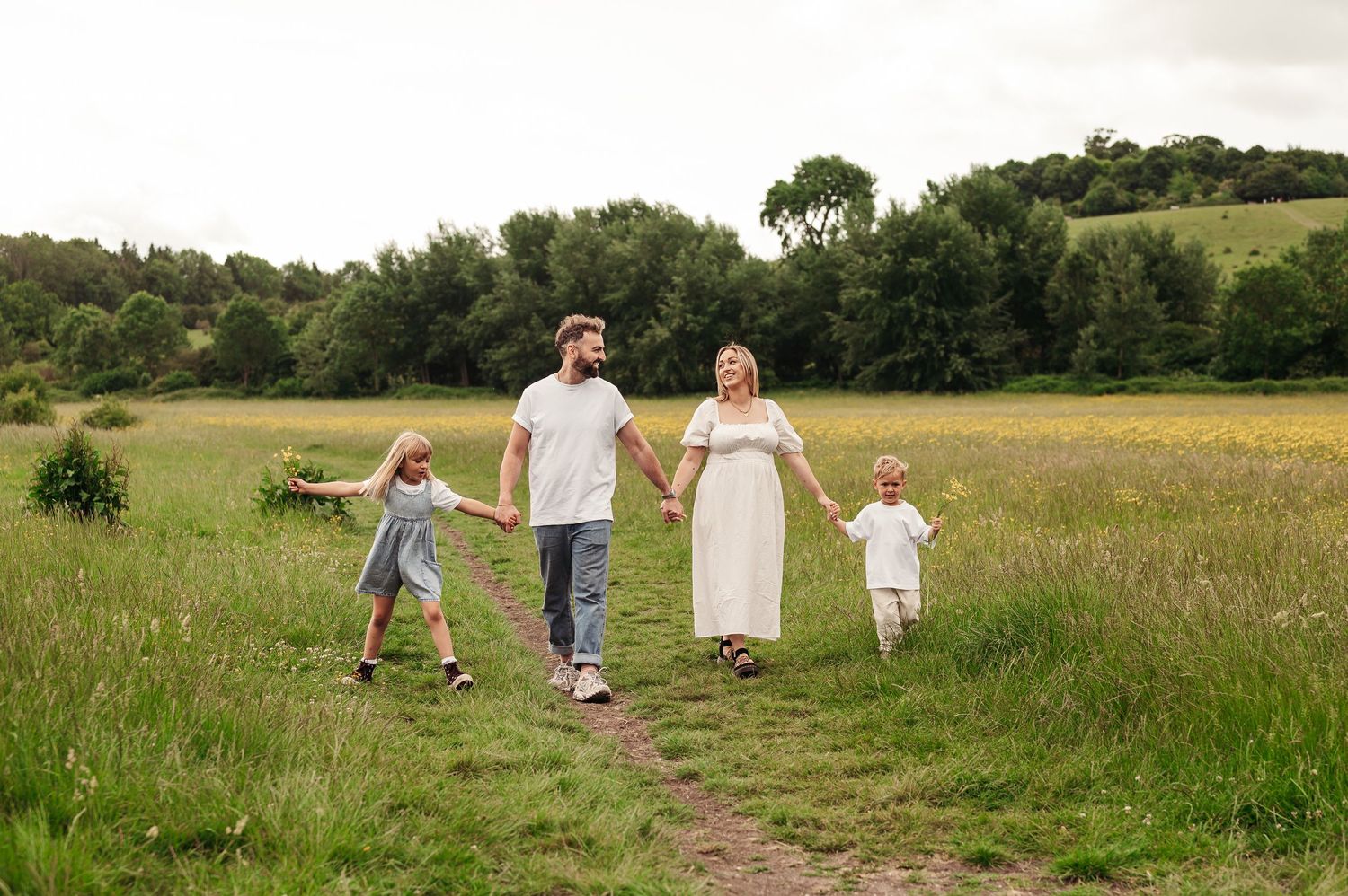 A family walks hand-in-hand along a dirt path through a sunny meadow surrounded by trees and rolling hills.