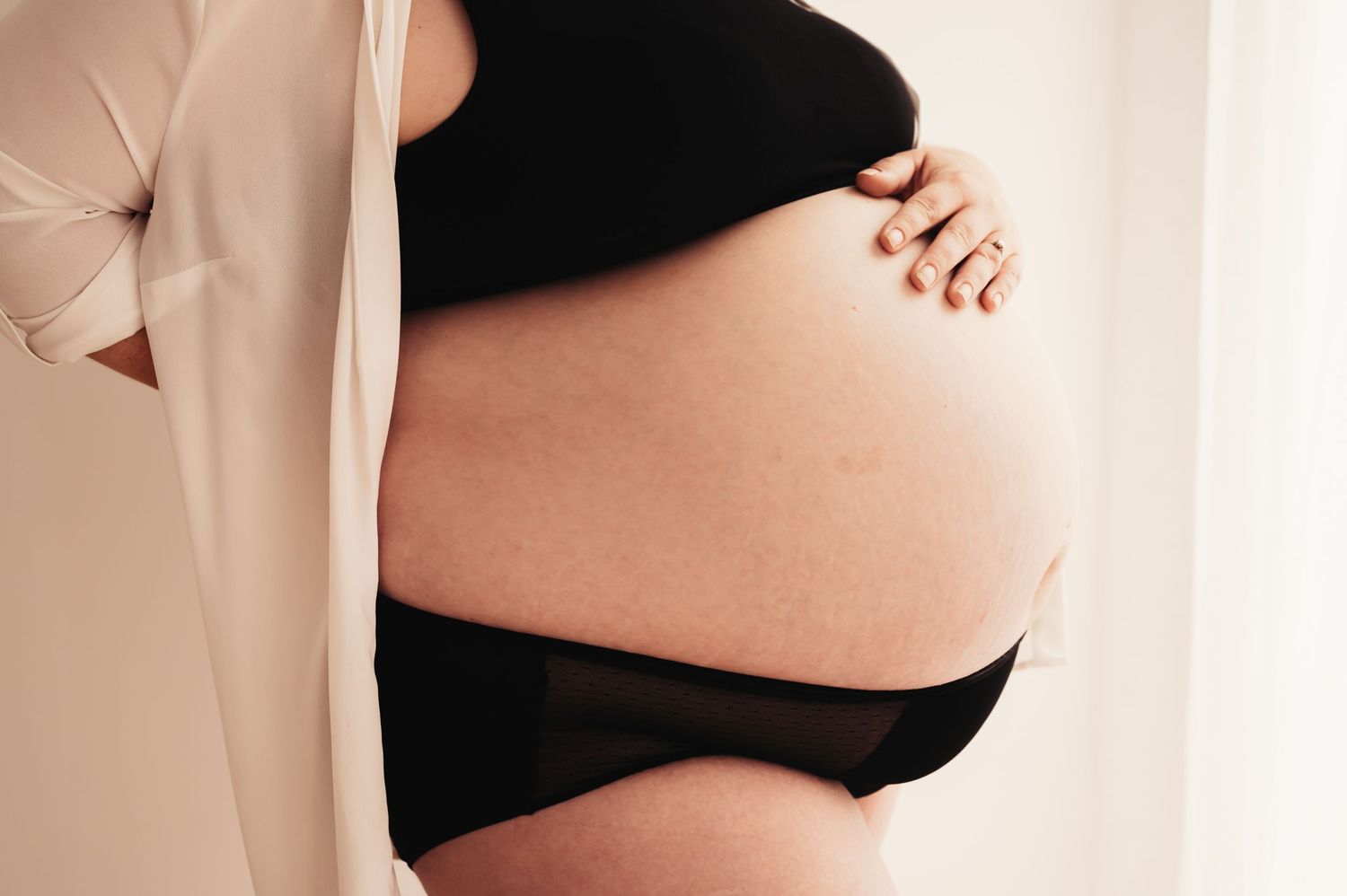 Close-up side view of pregnant belly in black undergarments against light background.