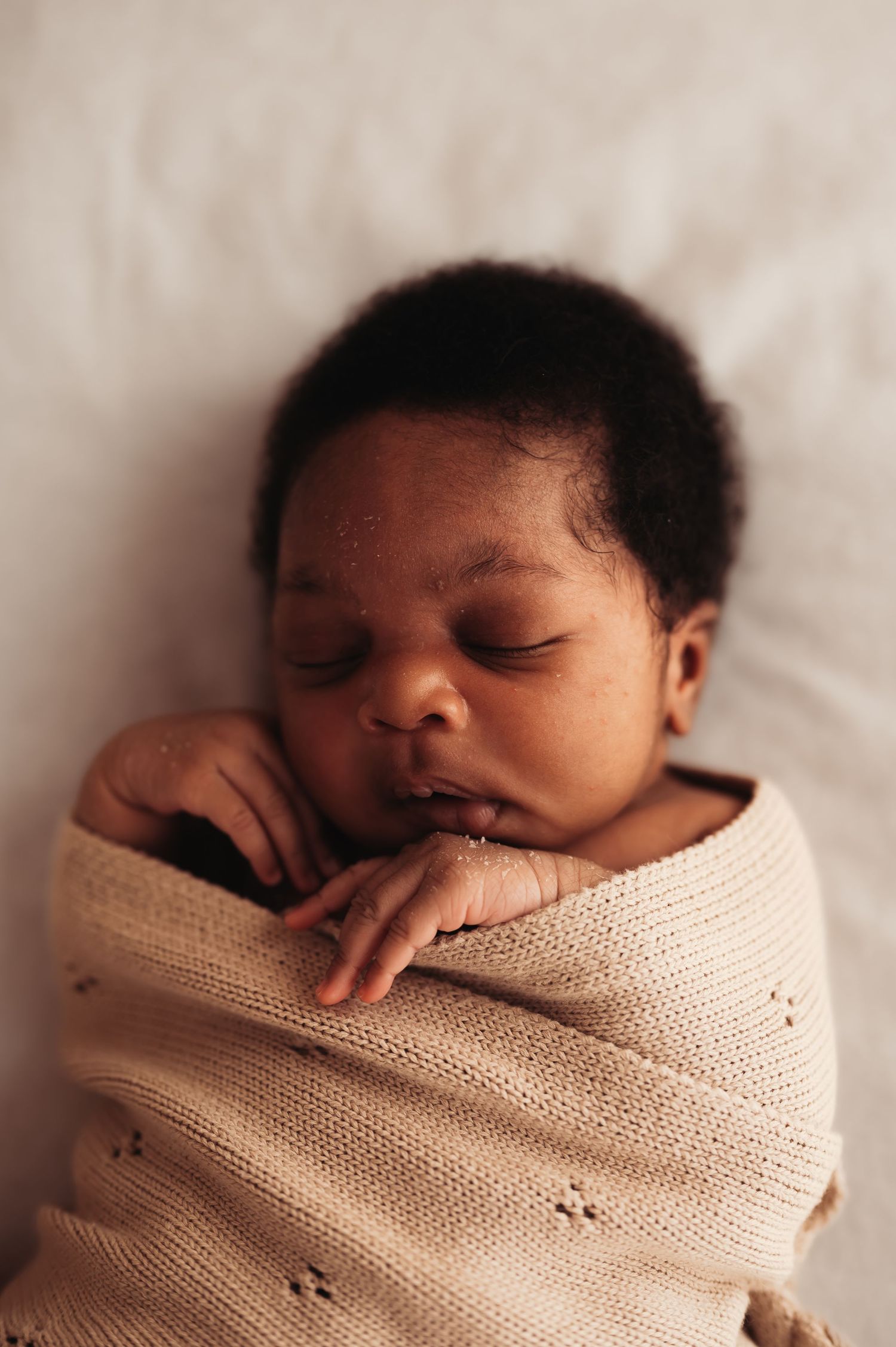 Peaceful newborn baby swaddled in a soft beige knit blanket, sleeping with tiny hands tucked under chin.
