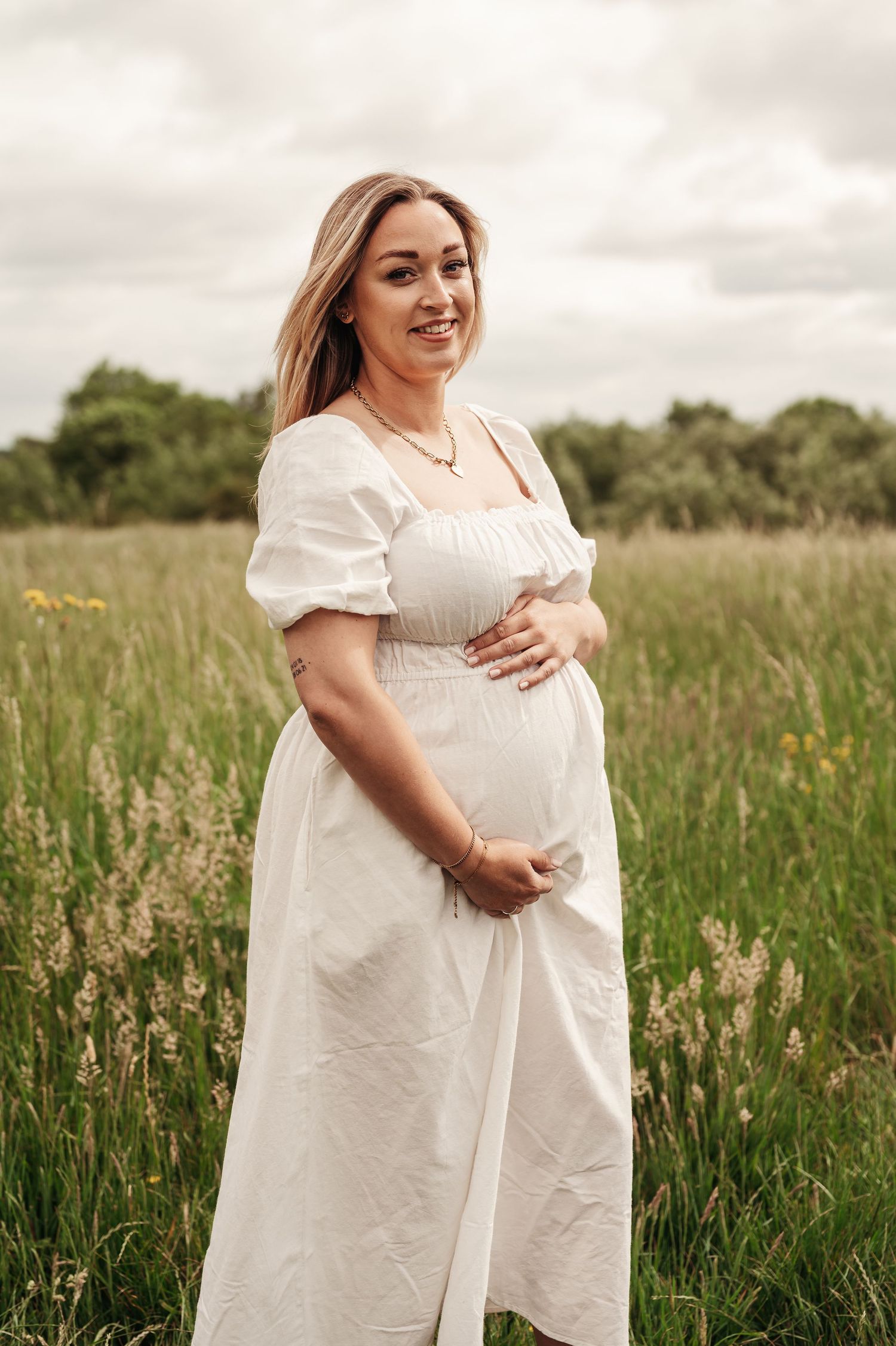 A person in a flowing white maternity dress poses in a serene meadow with tall grass and wildflowers.