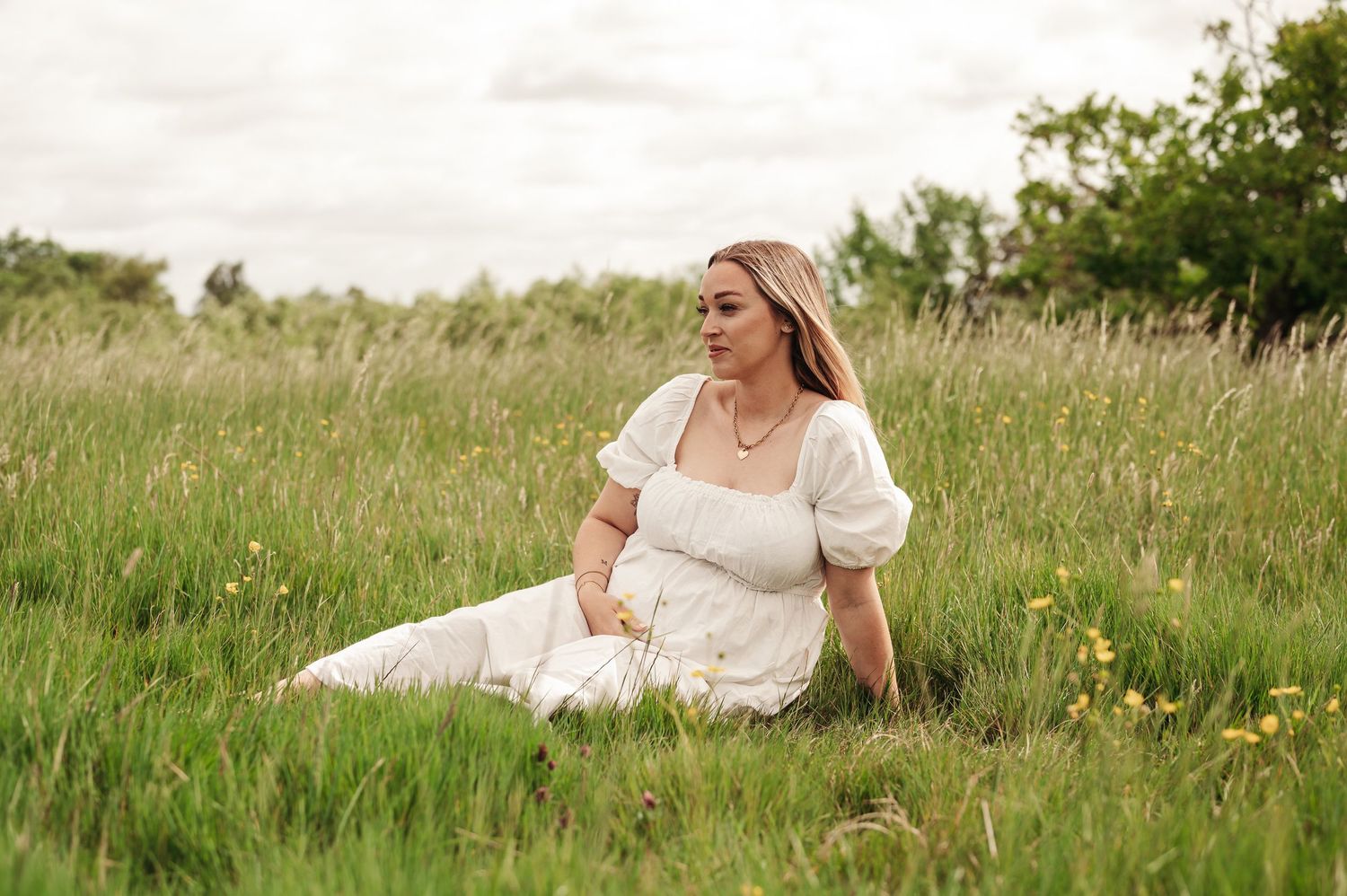 Someone in a white dress sits thoughtfully in a summer meadow with tall grass and wildflowers on a sunny day.