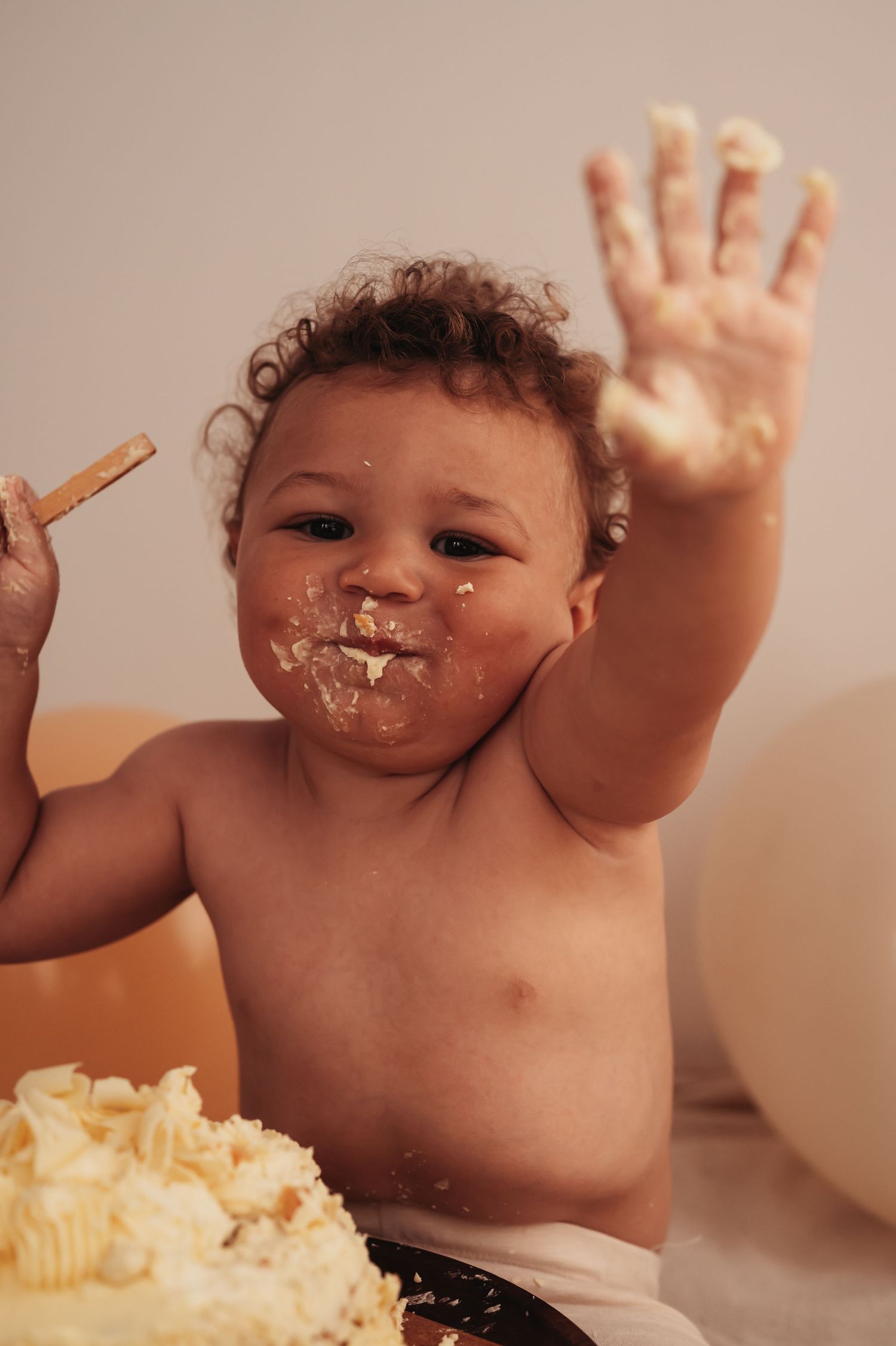 Messy baby covered in cake frosting waves during cake smash photo session.