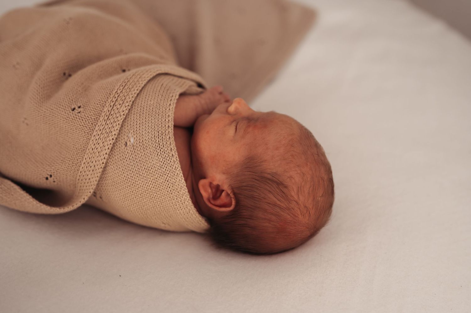Peaceful baby resting in cozy tan knitted wrap against light background.