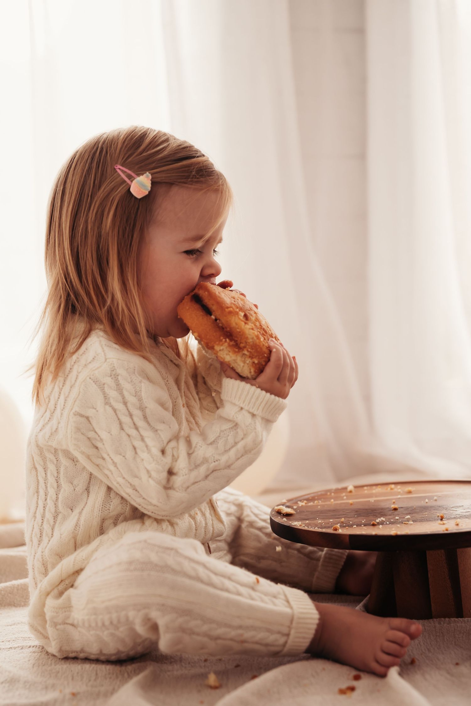A child in a knit sweater taking a big bite of cake while sitting on the floor.
