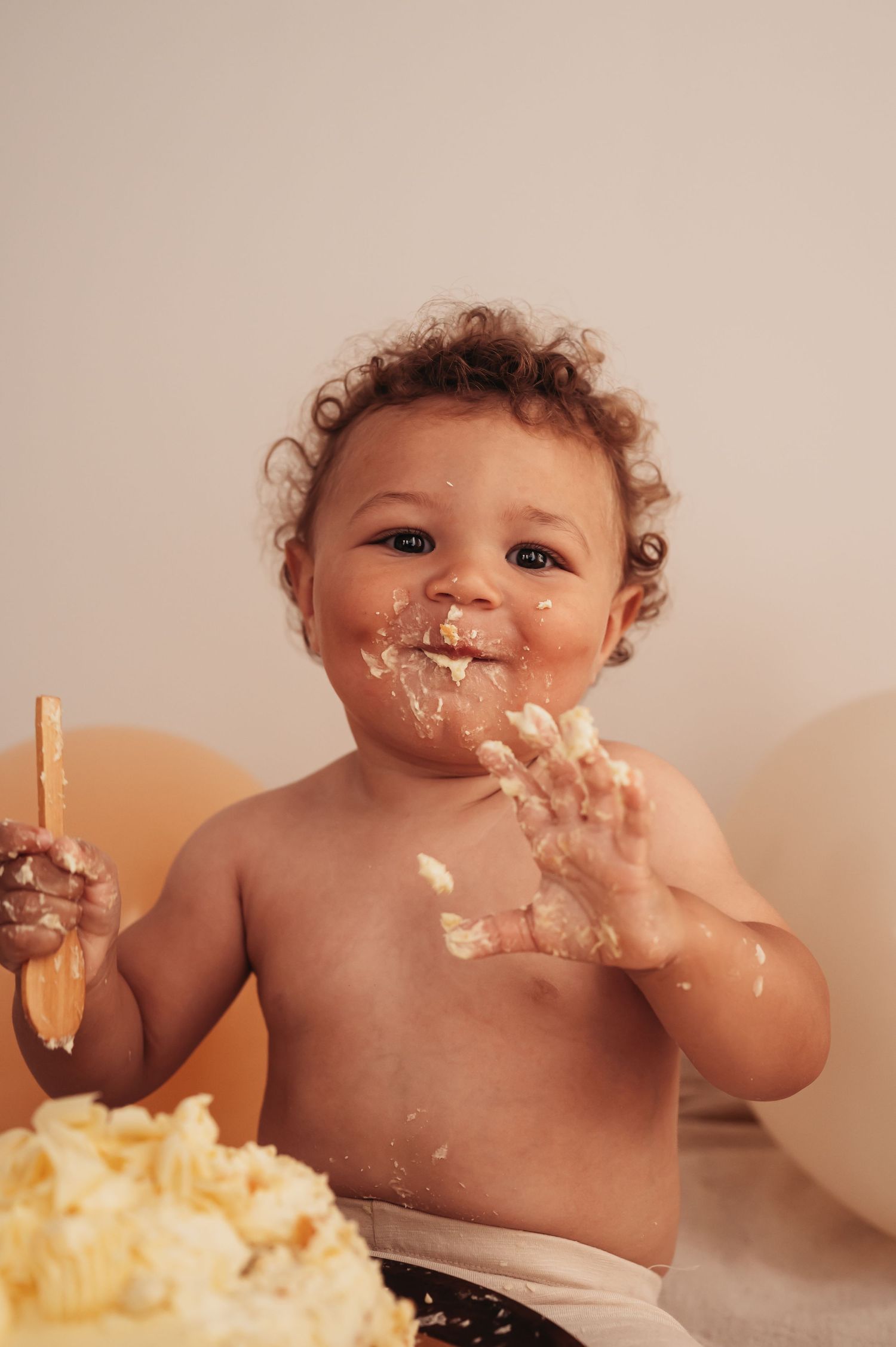 Messy baby playing with birthday cake and frosting while surrounded by peach balloons.
