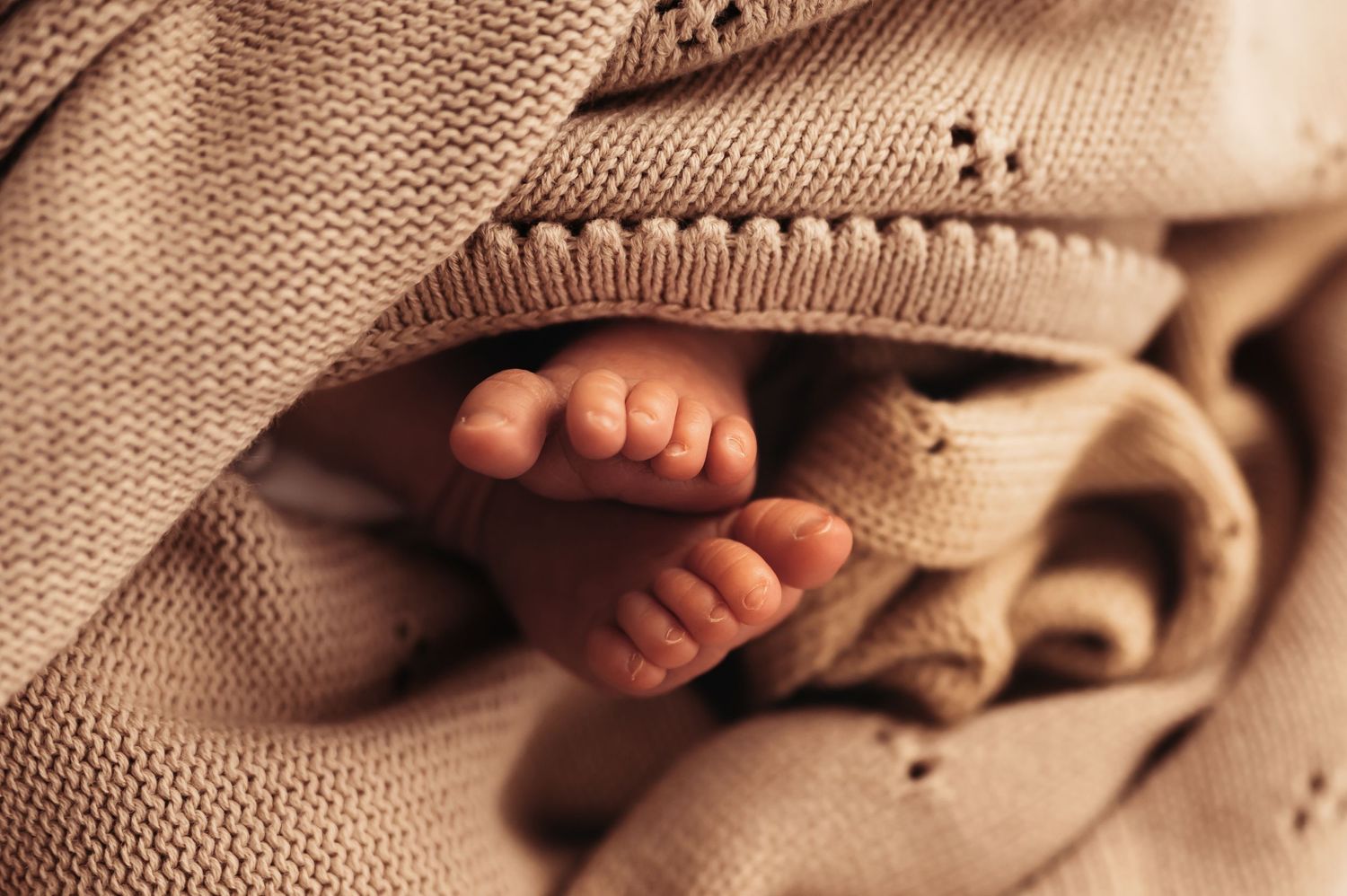 Tiny baby feet peek out from underneath a knitted beige blanket.