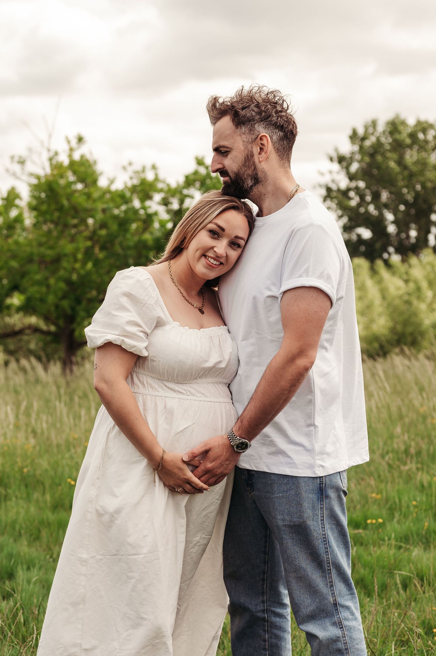 A couple shares a tender moment together in a grassy field, with her wearing a white dress and him in a casual t-shirt and jeans.