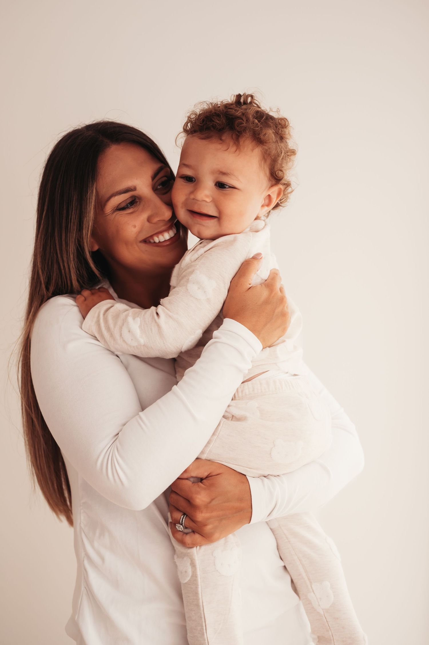 Mother and child sharing joyful embrace in matching white outfits against neutral background.