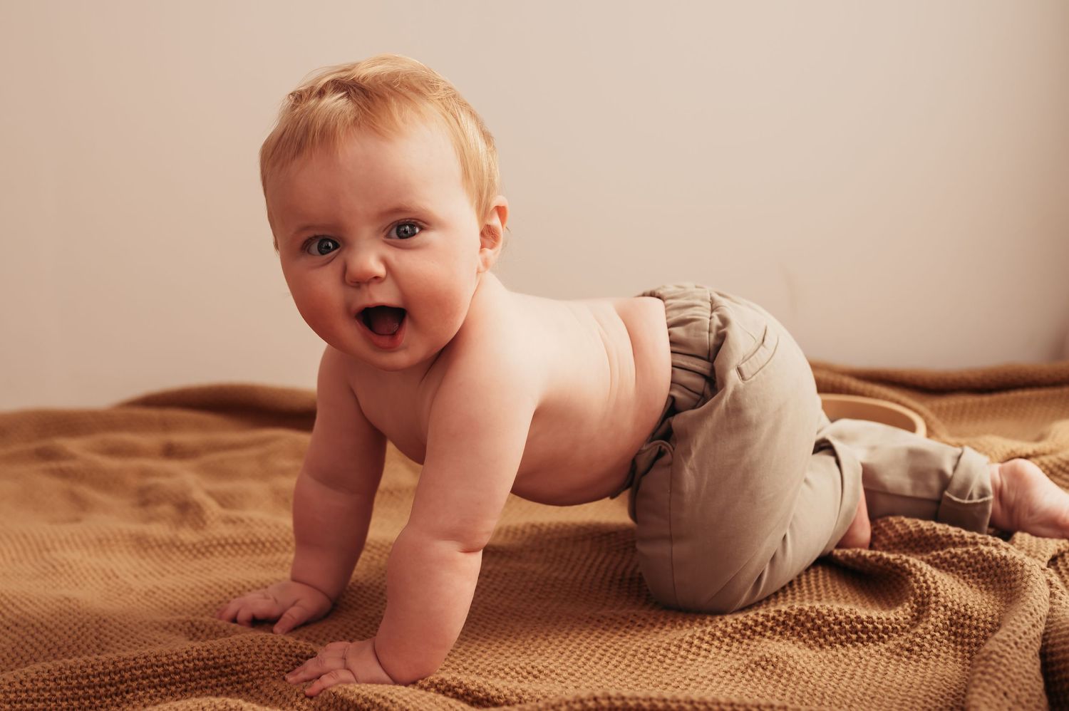 Happy baby crawling on a brown textured blanket, wearing tan bloomers, smiling with mouth open.