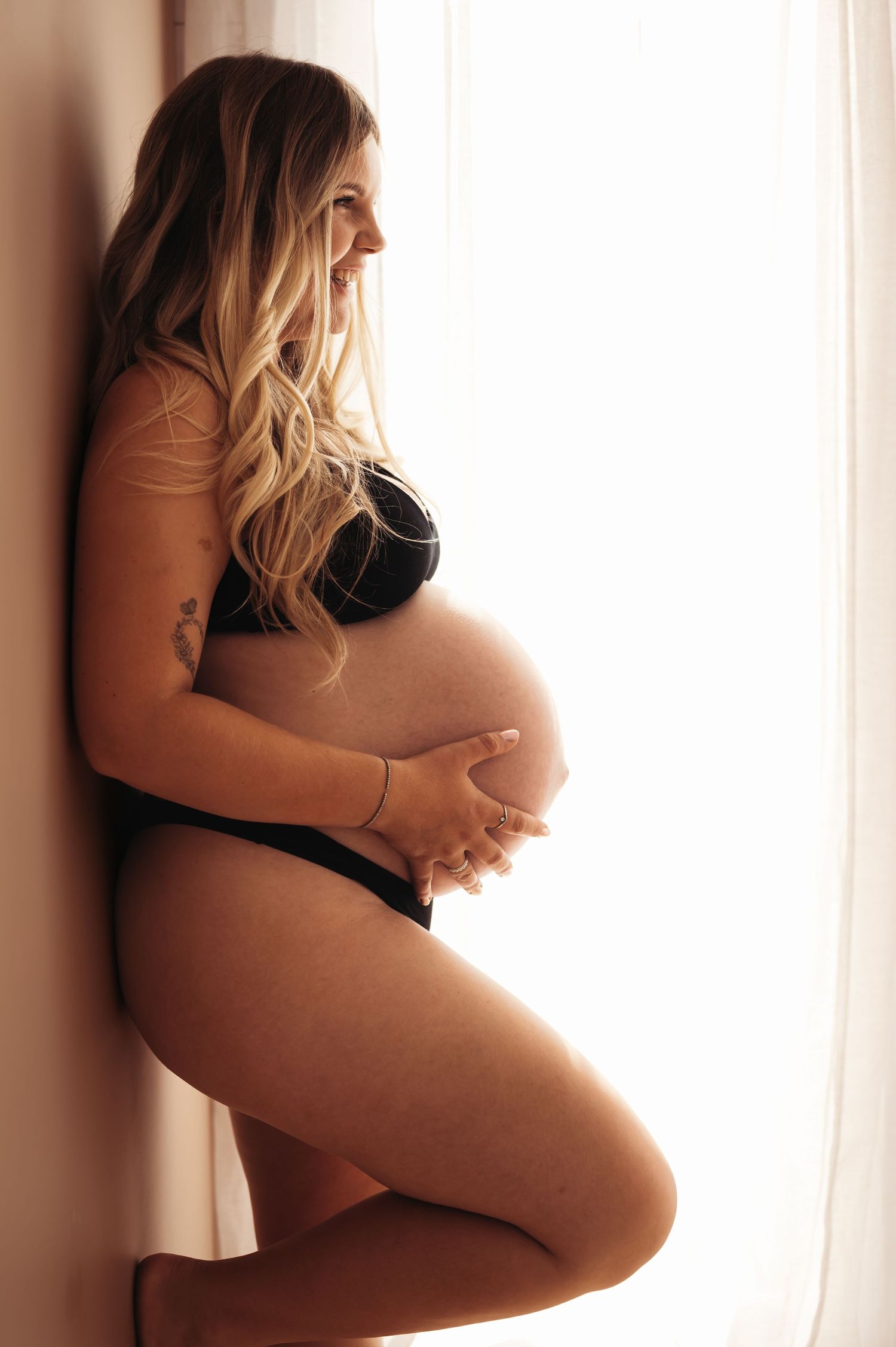 Pregnant woman in black lingerie cradling her baby bump while leaning against a wall near a bright window.