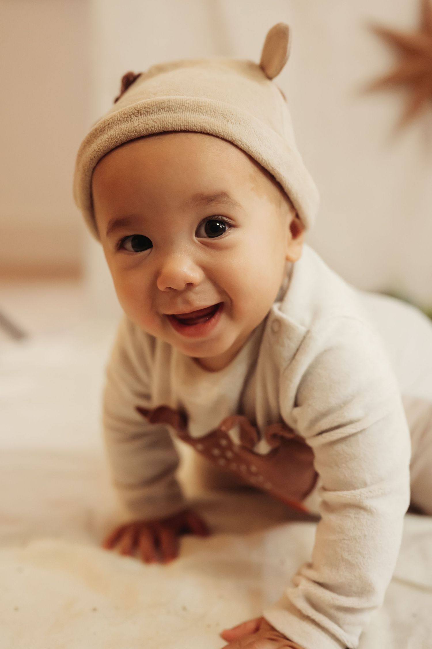 Adorable smiling baby in a beige deer outfit and bear ear hat, crawling on a soft surface indoors.