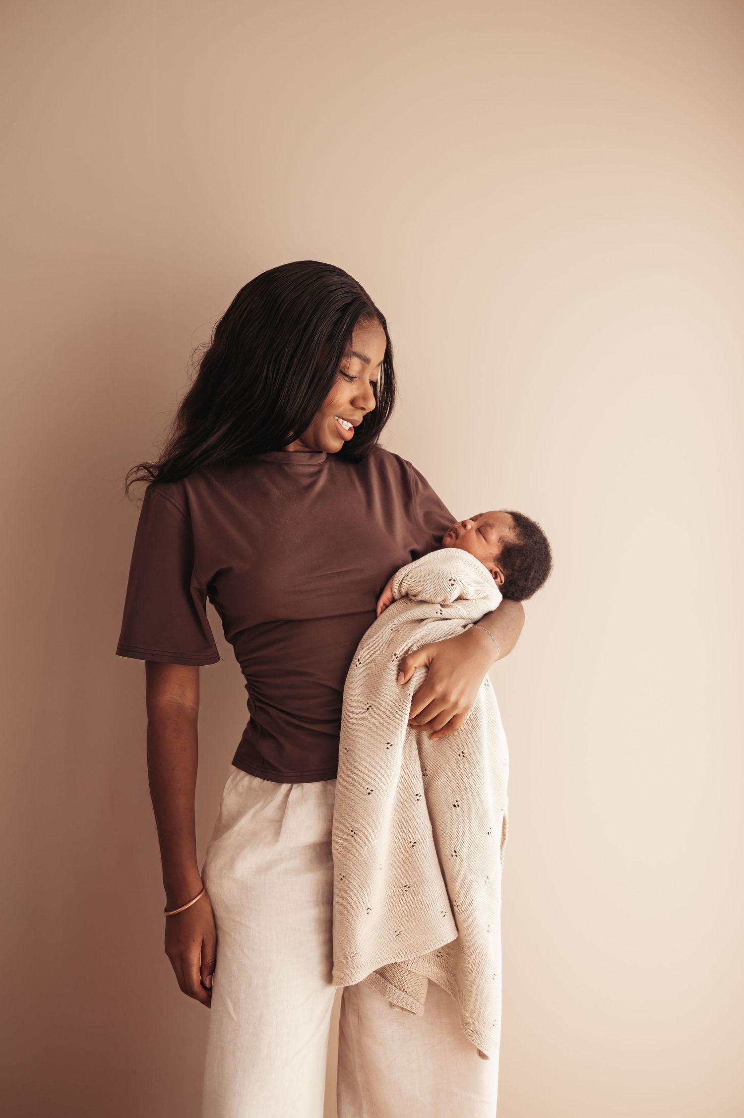 A young Black mother in a brown top holds her sleeping newborn wrapped in a light patterned blanket.