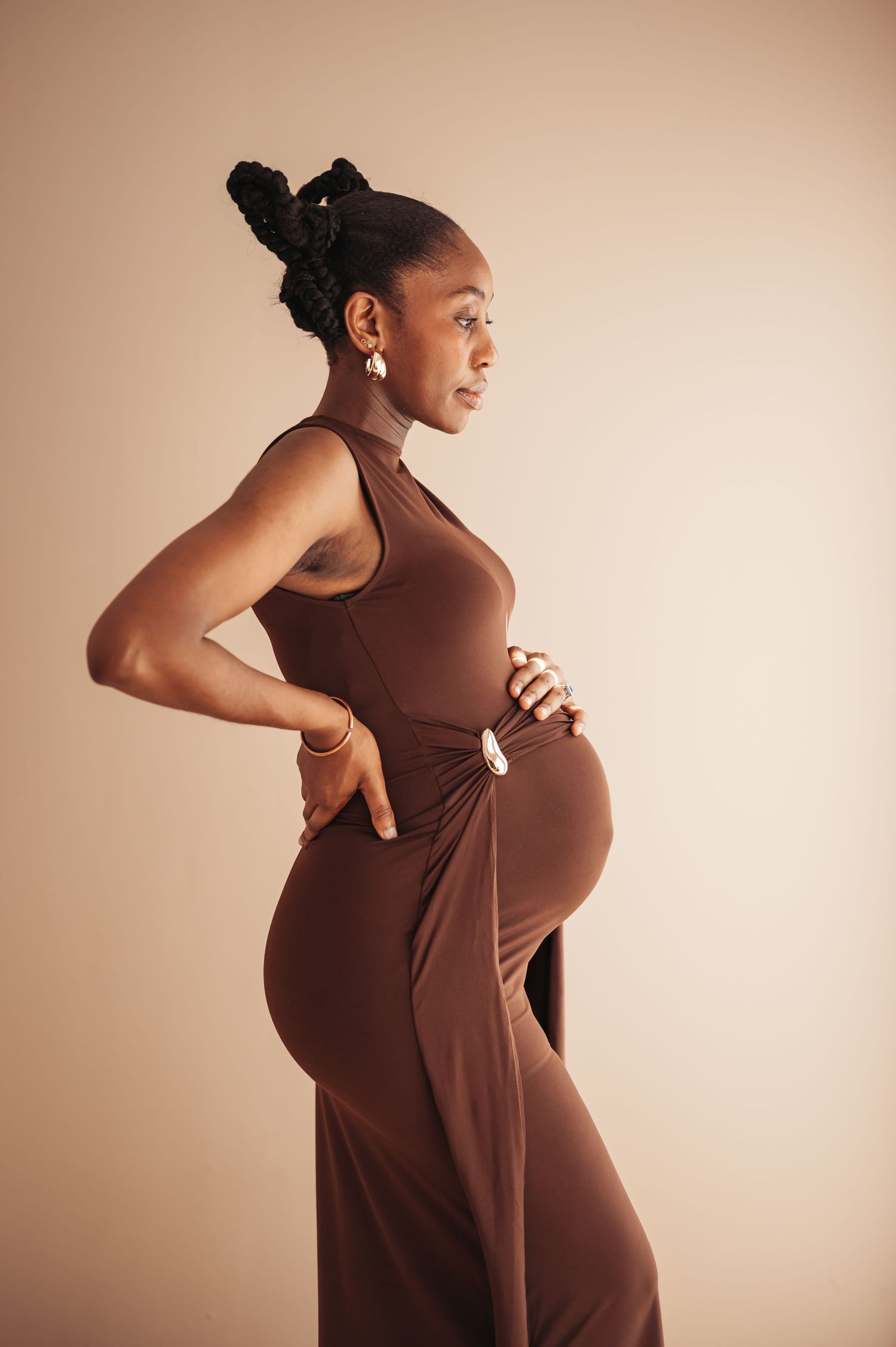 Pregnant Black woman in brown sleeveless maxi dress posing sideways, hands on hips, hair up, wearing gold earrings.