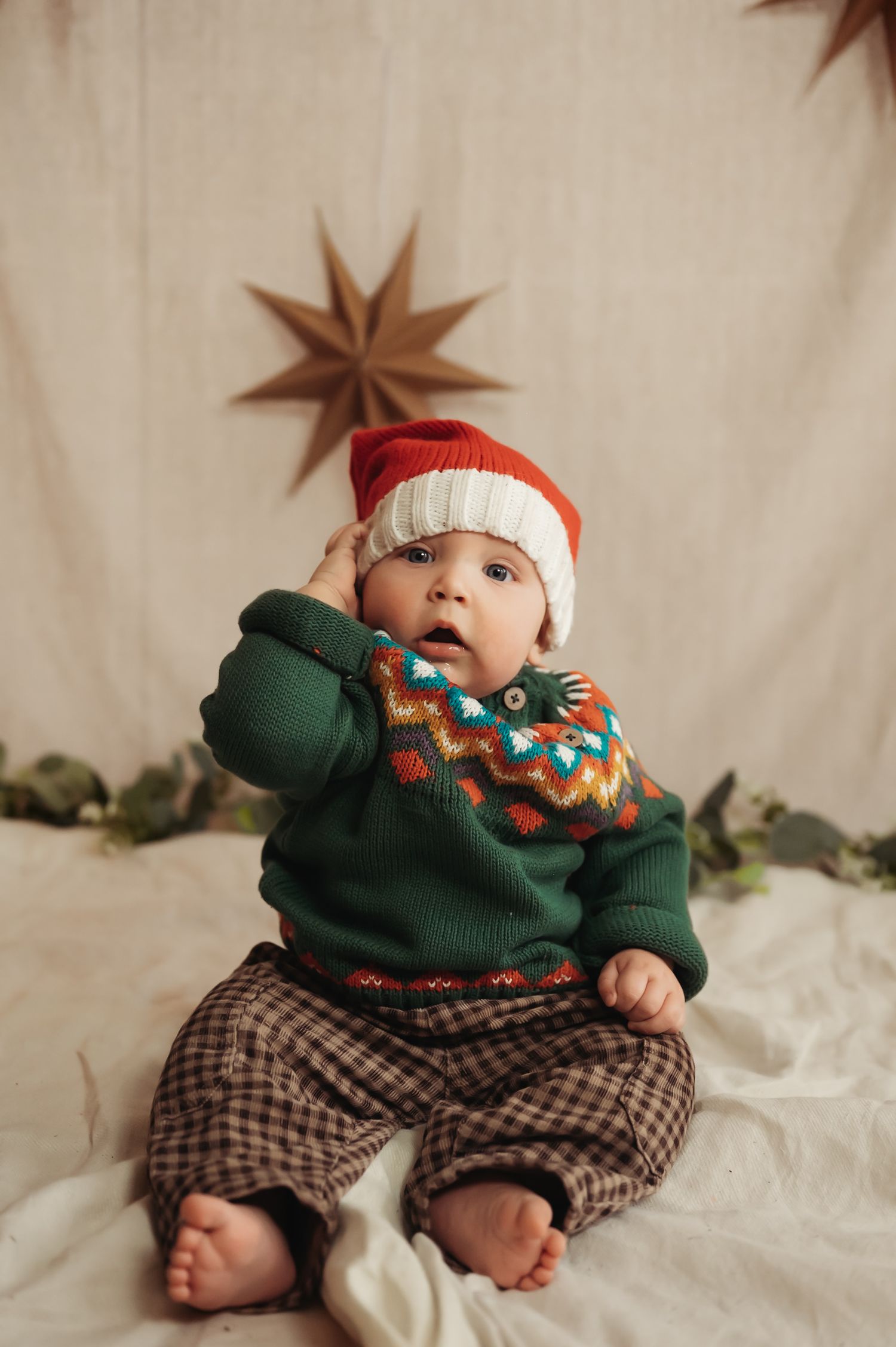 Baby in Santa hat and green Nordic sweater sits barefoot before star decor, perfect Christmas portrait.